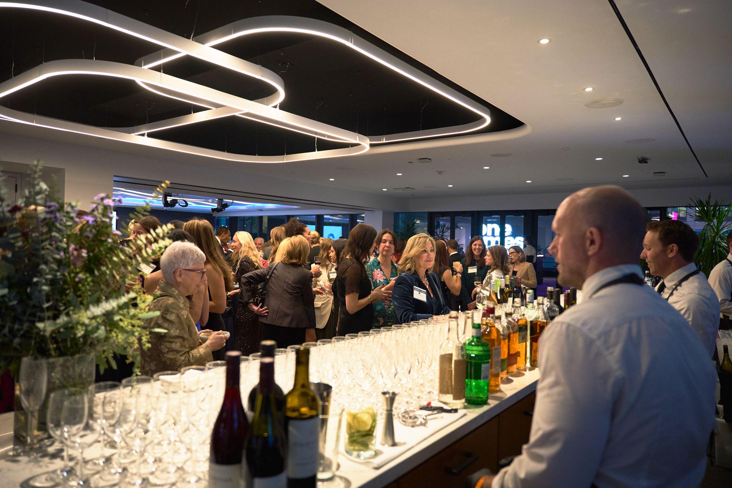 Guests lining a long lit bar stocked with wine and cocktails at an evening corporate party — Business Parties photography, Austin Guests lining a long lit bar stocked with wine and cocktails at an evening corporate party.