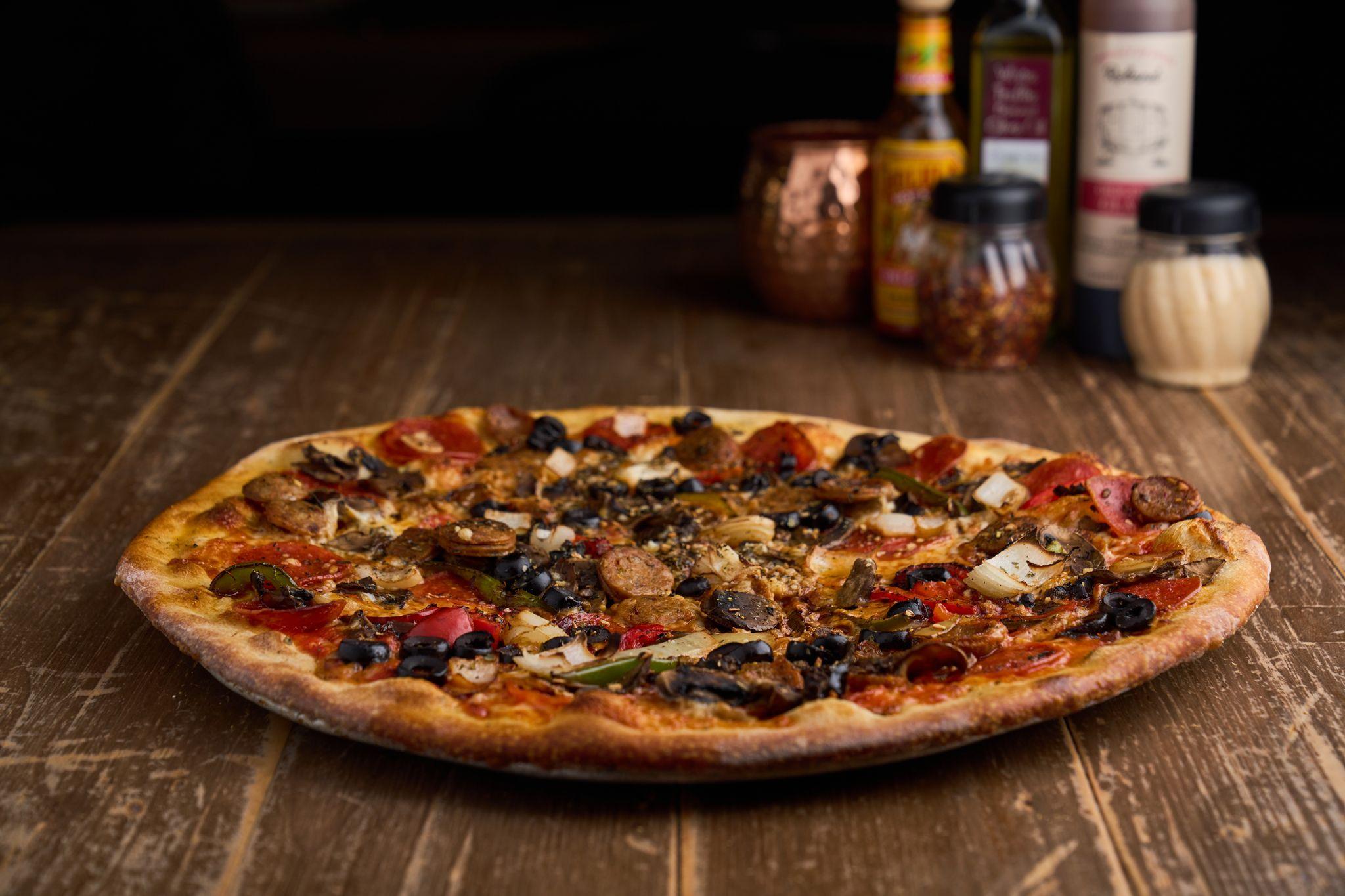 Pizza on a dark wooden table in low light, copper mugs behind it — Food & restaurant photography, Austin A pizza on a dark wooden table in low light, copper mugs behind it.