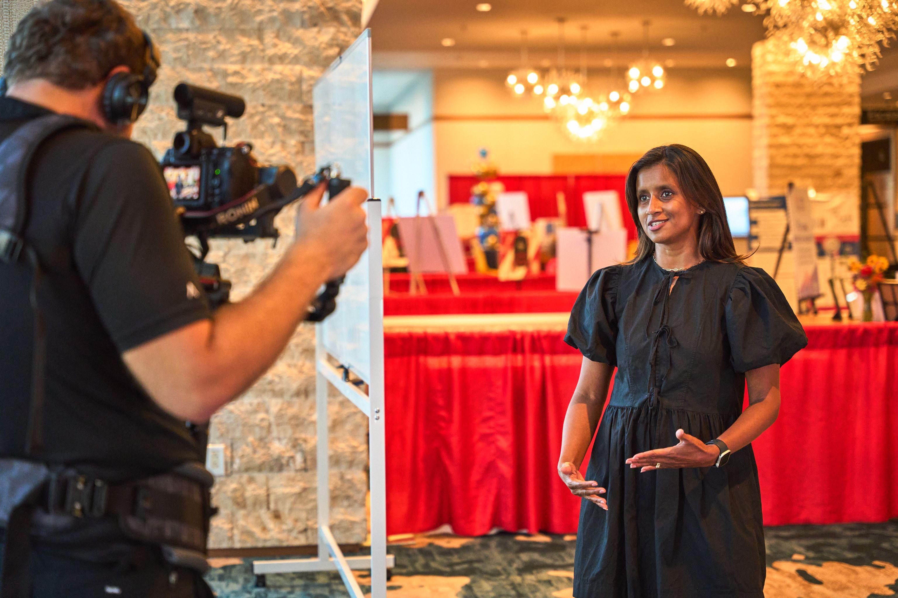 Camera operator filming a woman being interviewed beside a red-draped display table at an event — Production behind-the-scenes, Austin Camera operator filming a woman being interviewed beside a red-draped display table at an event