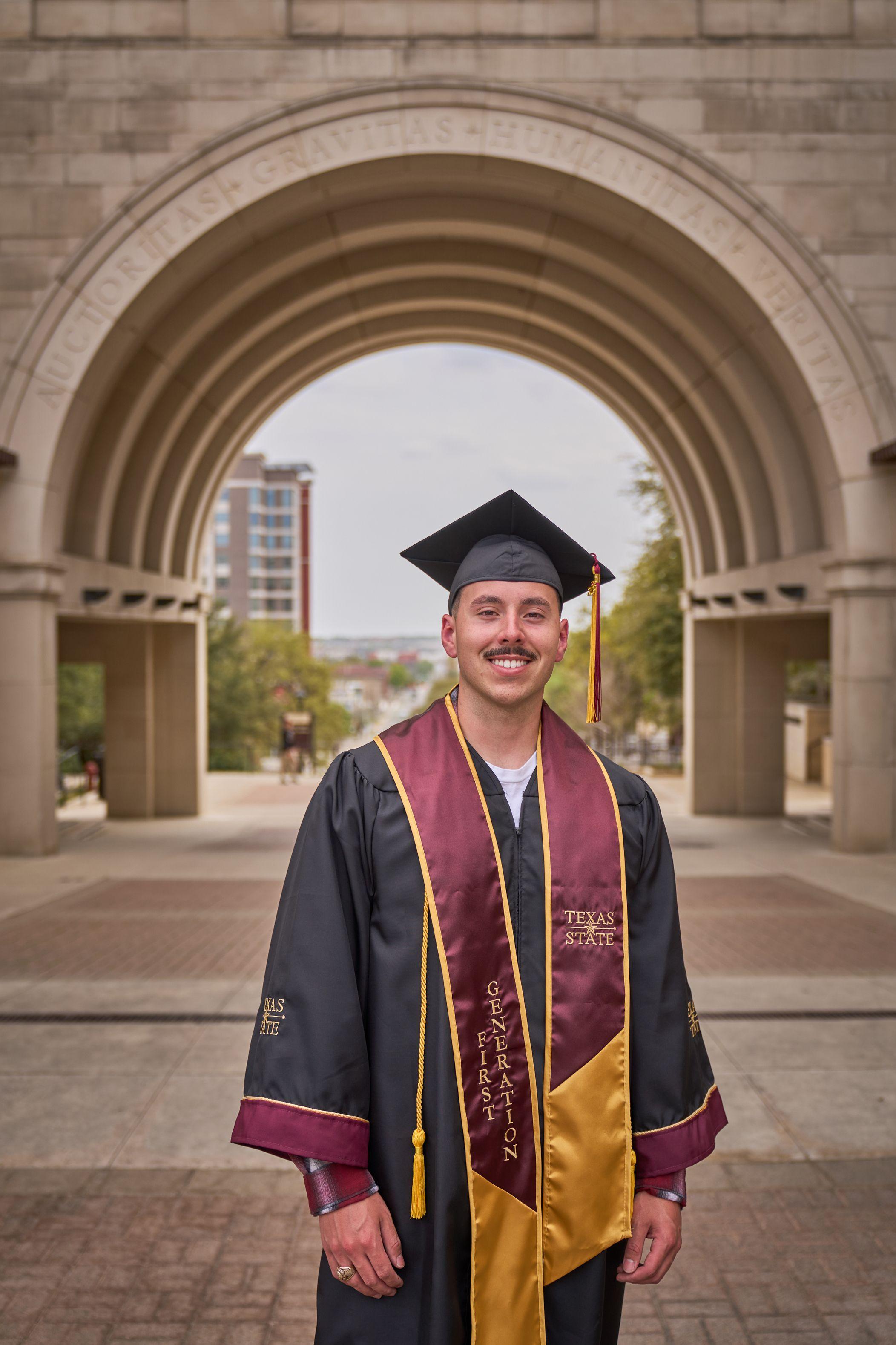 Graduate in cap, gown and maroon stole standing beneath the Texas State arch — Texas State University photography, Austin A graduate in cap, gown and maroon stole standing beneath the Texas State arch