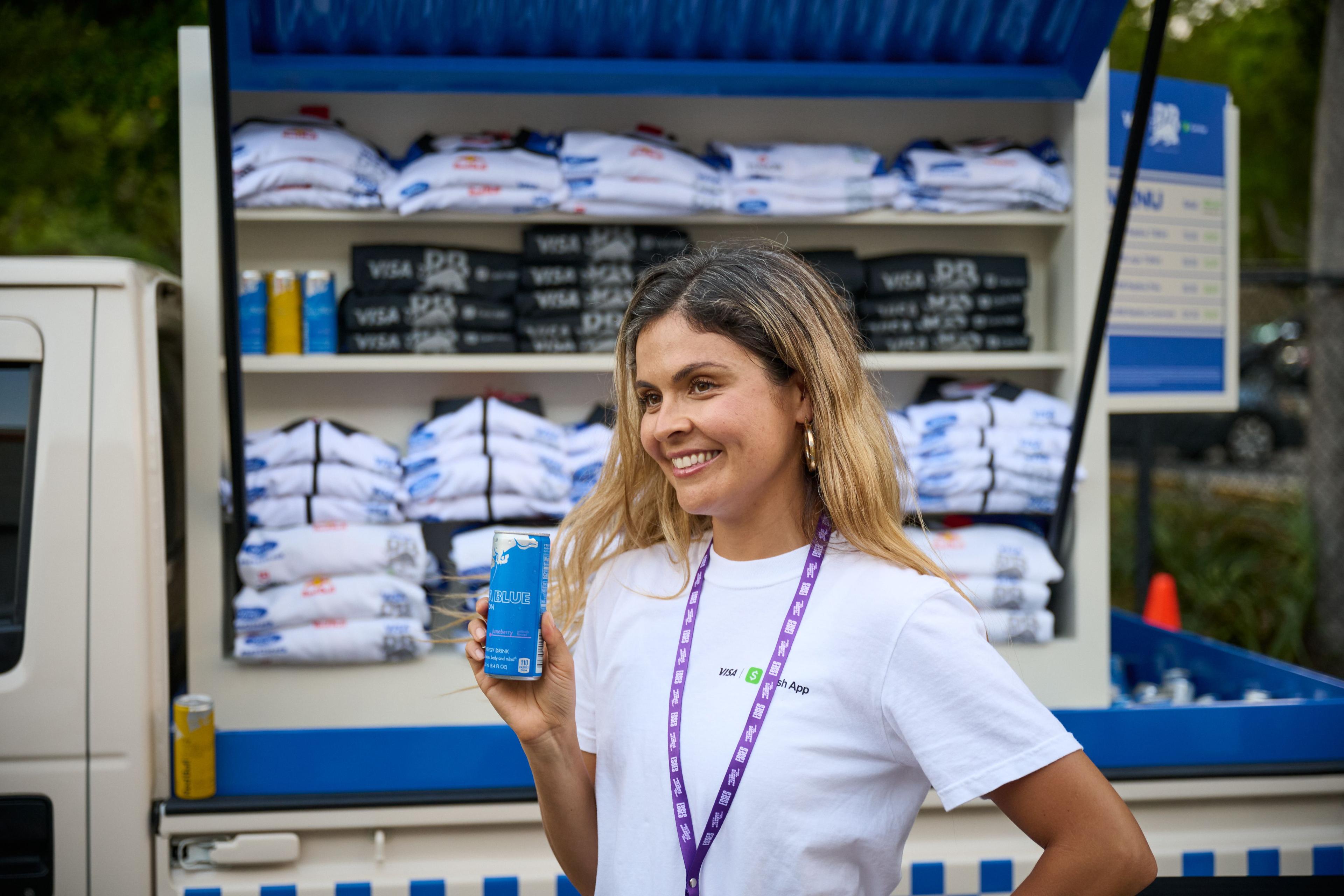 Brand host in a white shirt holding out a blue can in front of shelves of folded event merch — Brand Collaborations photography, Austin A brand host in a white shirt holding out a blue can in front of shelves of folded event merch.
