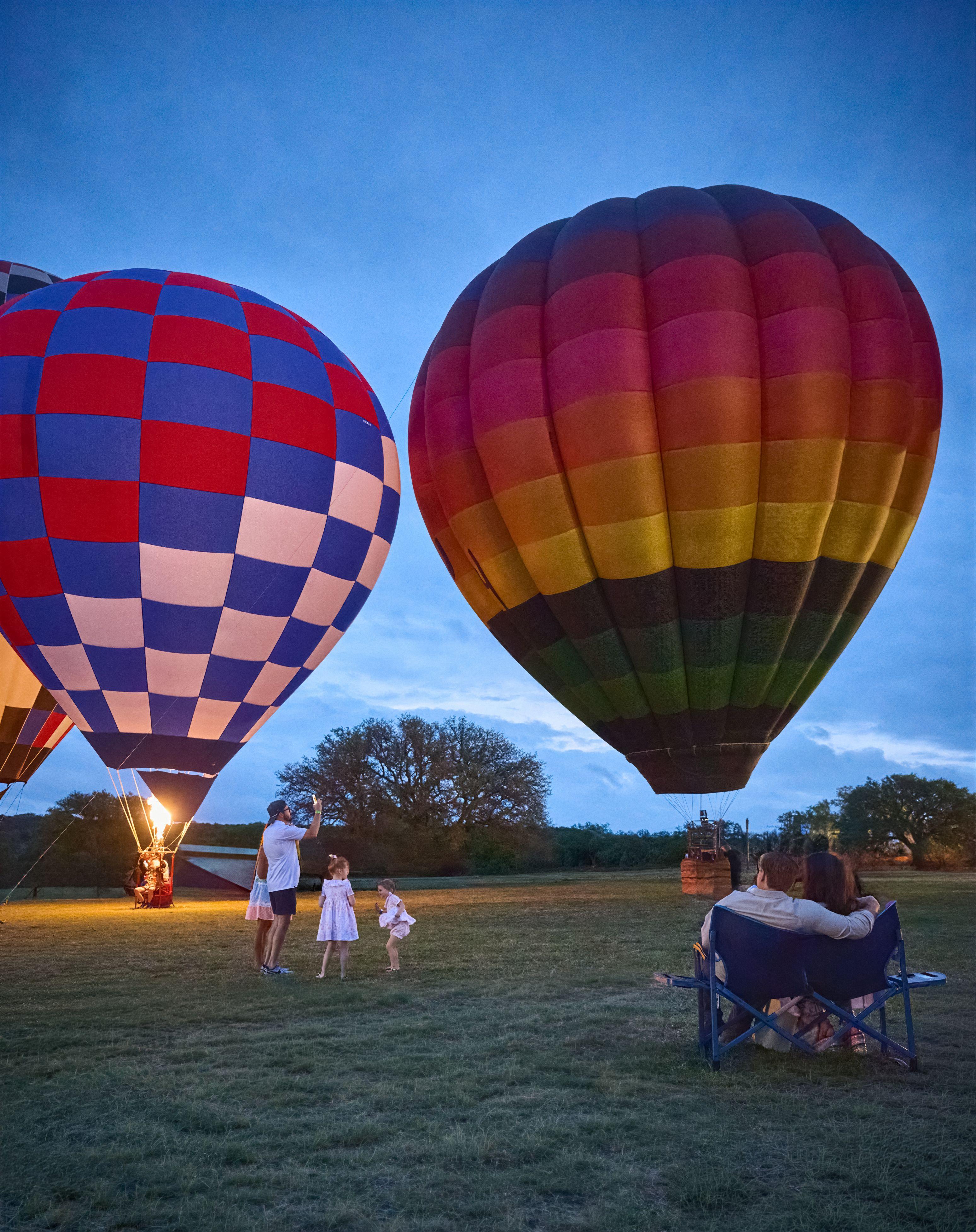 Two hot air balloons inflated on a field at dusk with families watching — Hospitality & leisure photography, Austin Two hot air balloons inflated on a field at dusk with families watching.