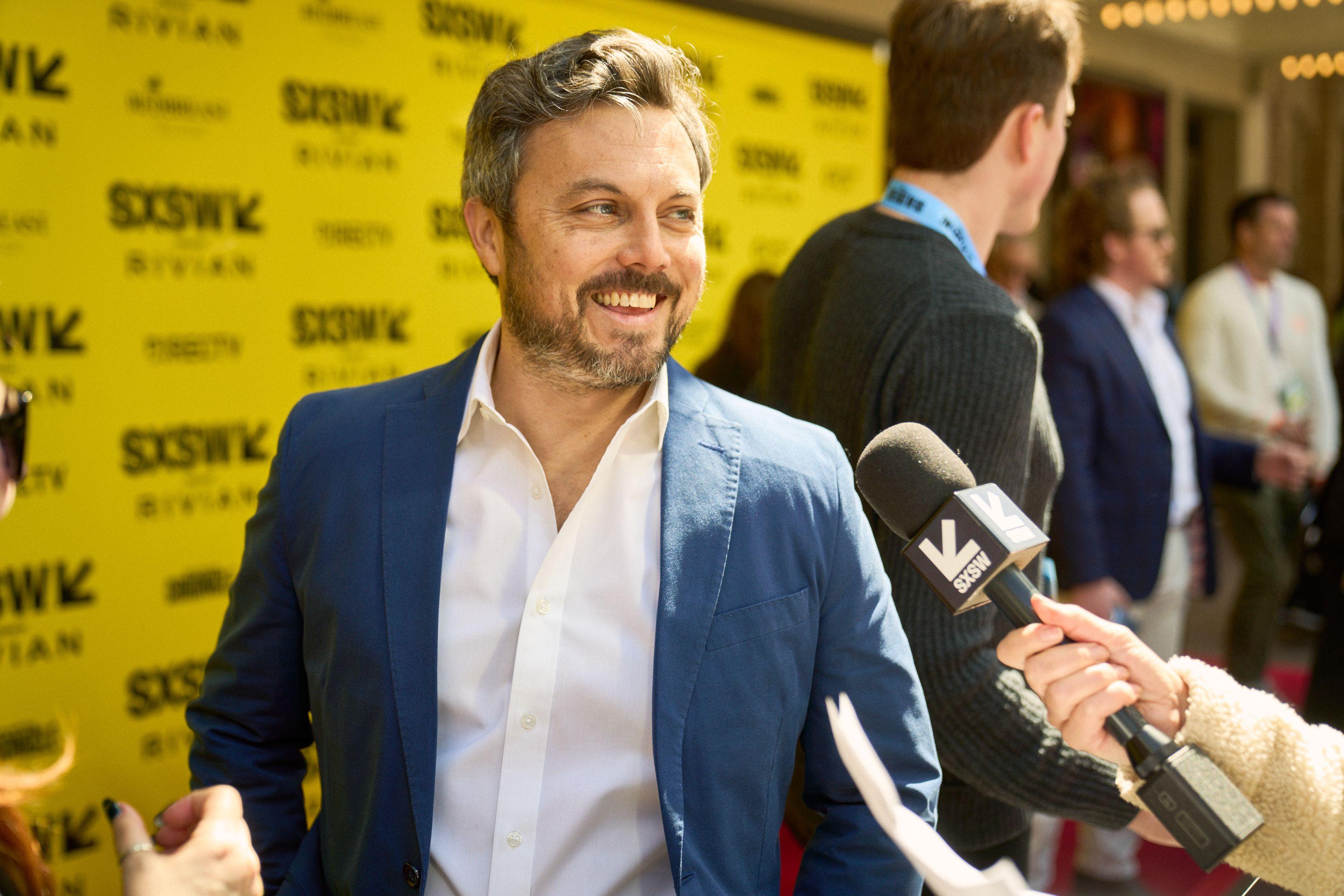 Man in a blue blazer smiling on the red carpet as reporters gather at the edge of frame — Production behind-the-scenes, Austin Man in a blue blazer smiling on the red carpet as reporters gather at the edge of frame