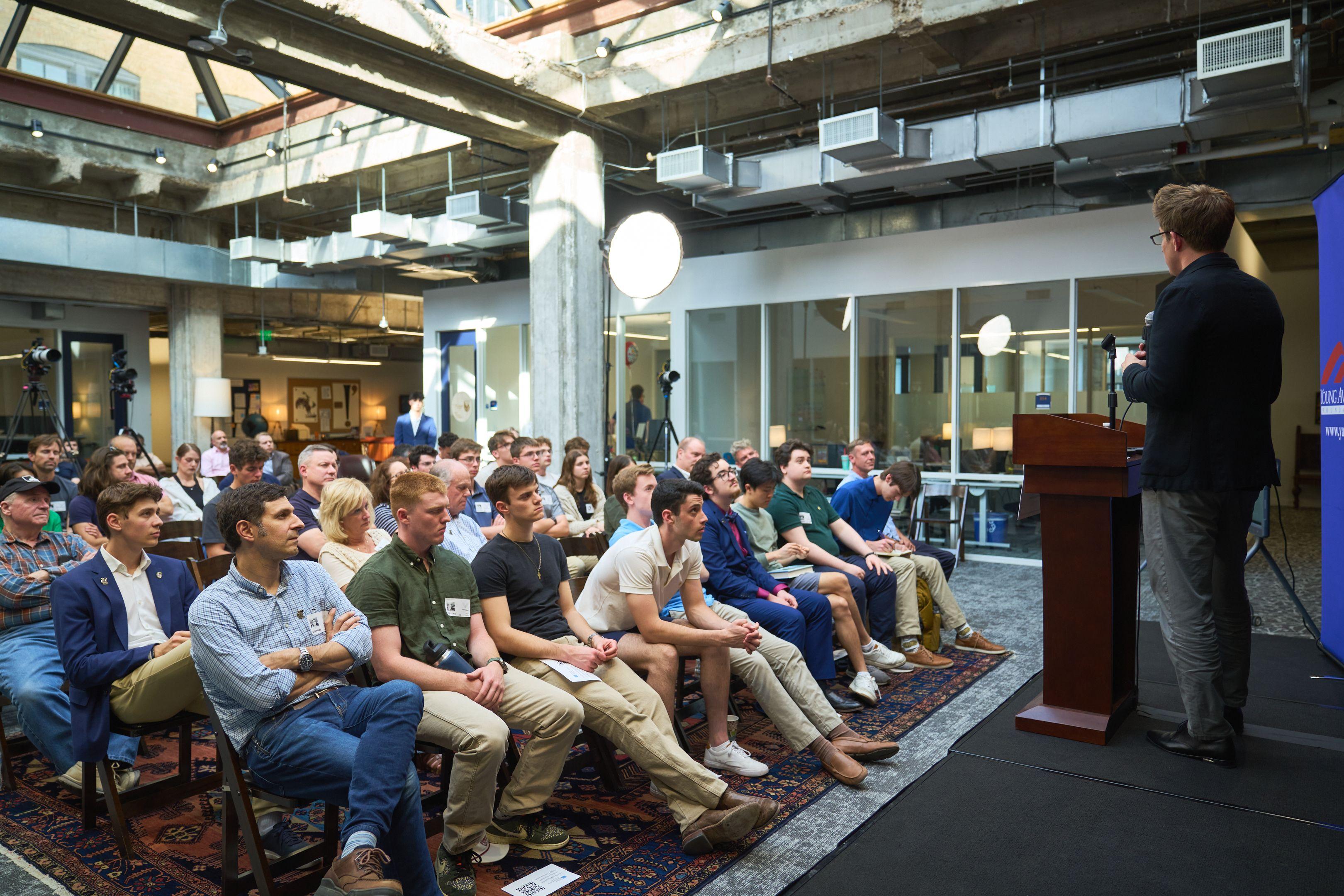 Audience seated on folding chairs in a high-ceilinged industrial room listening to a talk — Production behind-the-scenes, Austin Audience seated on folding chairs in a high-ceilinged industrial room listening to a talk