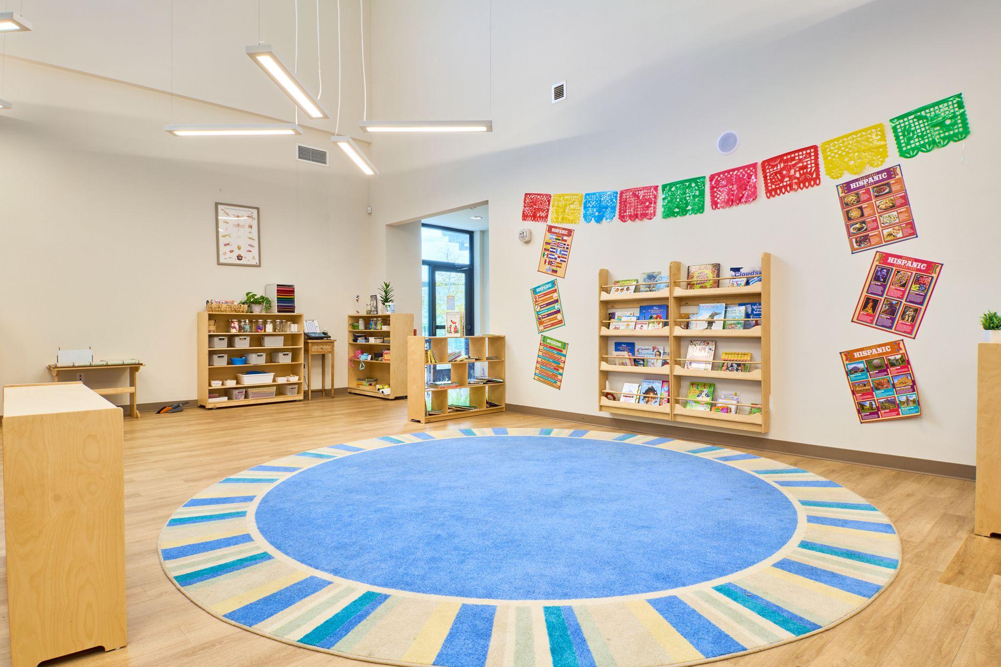 Empty early-years classroom with a blue and yellow circular rug, low bookshelves — School event photography, Austin Empty early-years classroom with a blue and yellow circular rug, low bookshelves and papel picado bunting