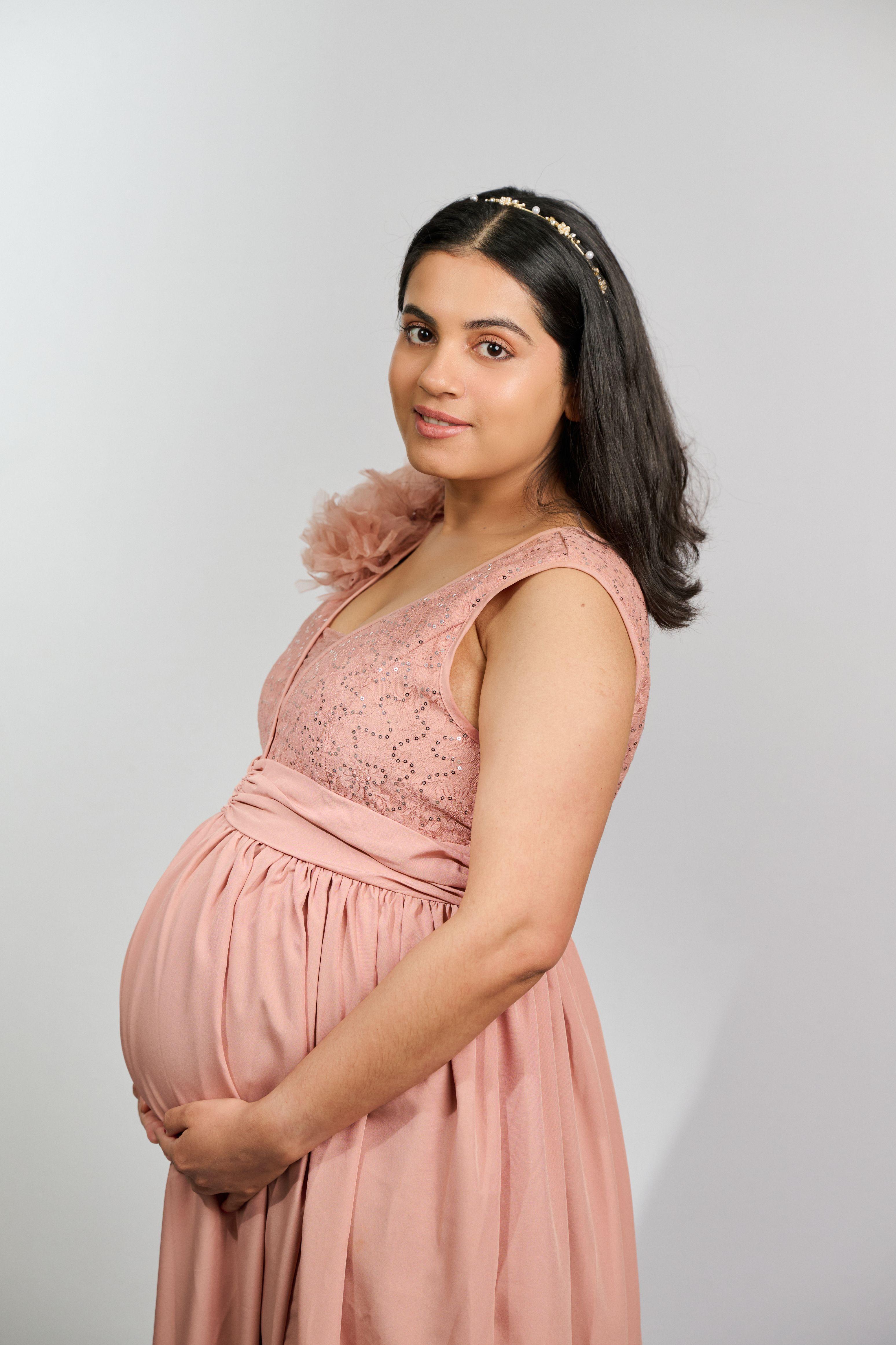 Pregnant woman in a pink dress with both hands under her bump against a light studio background — Commercial photo studio, Austin Pregnant woman in a pink dress with both hands under her bump against a light studio background