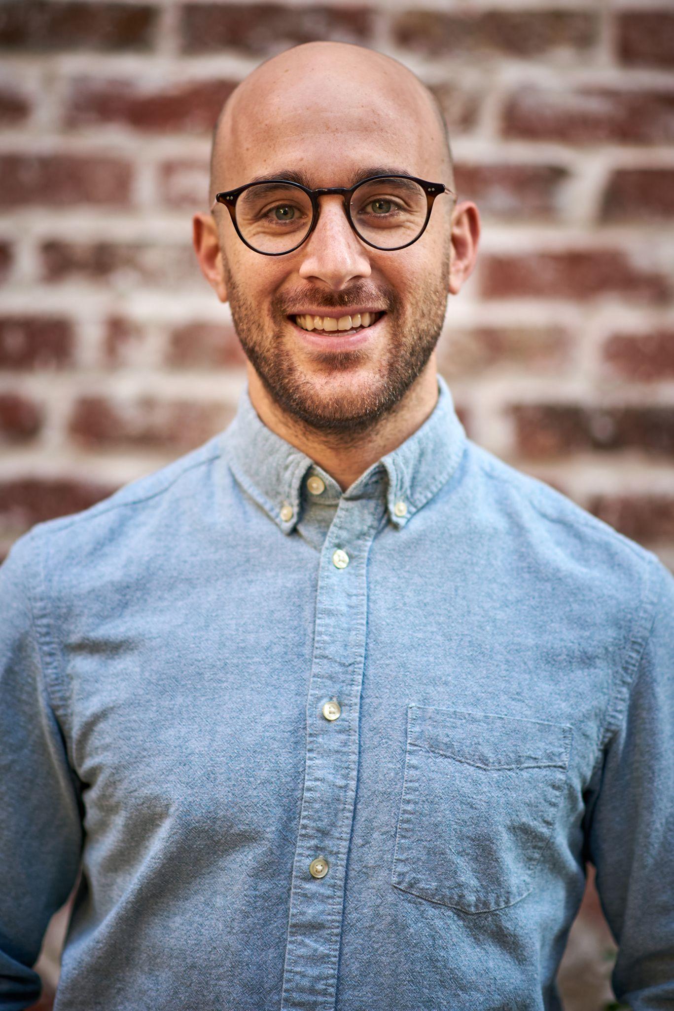 Man in glasses and a light blue shirt smiling in front of an exposed brick wall — Business headshots, Austin A man in glasses and a light blue shirt smiling in front of an exposed brick wall