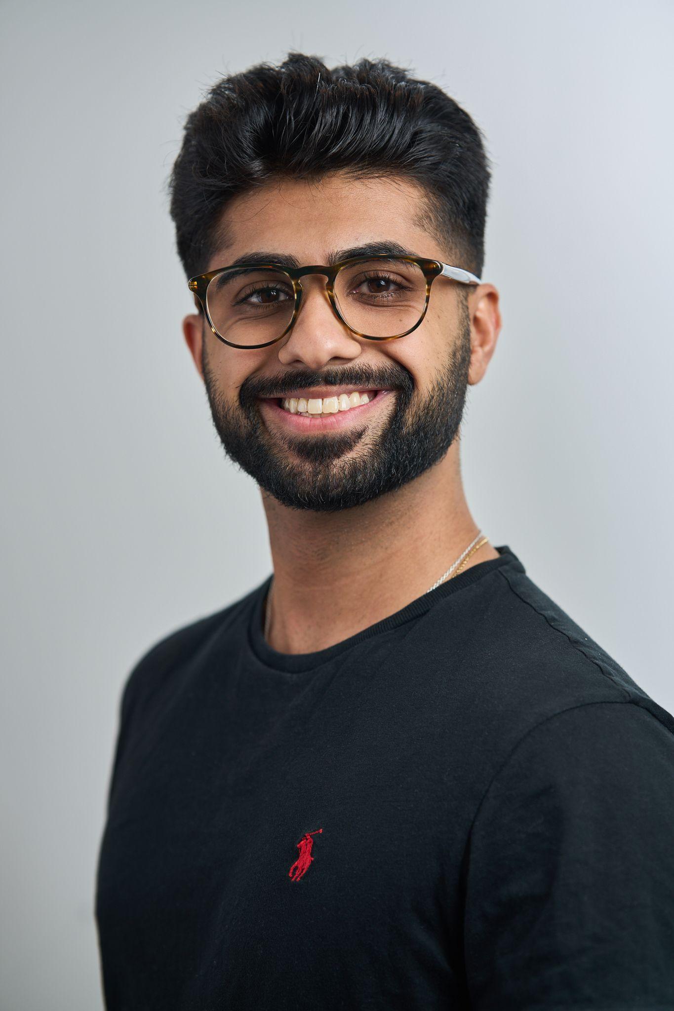 Young man in glasses and a black polo shirt smiling against a plain light background — Business headshots, Austin A young man in glasses and a black polo shirt smiling against a plain light background
