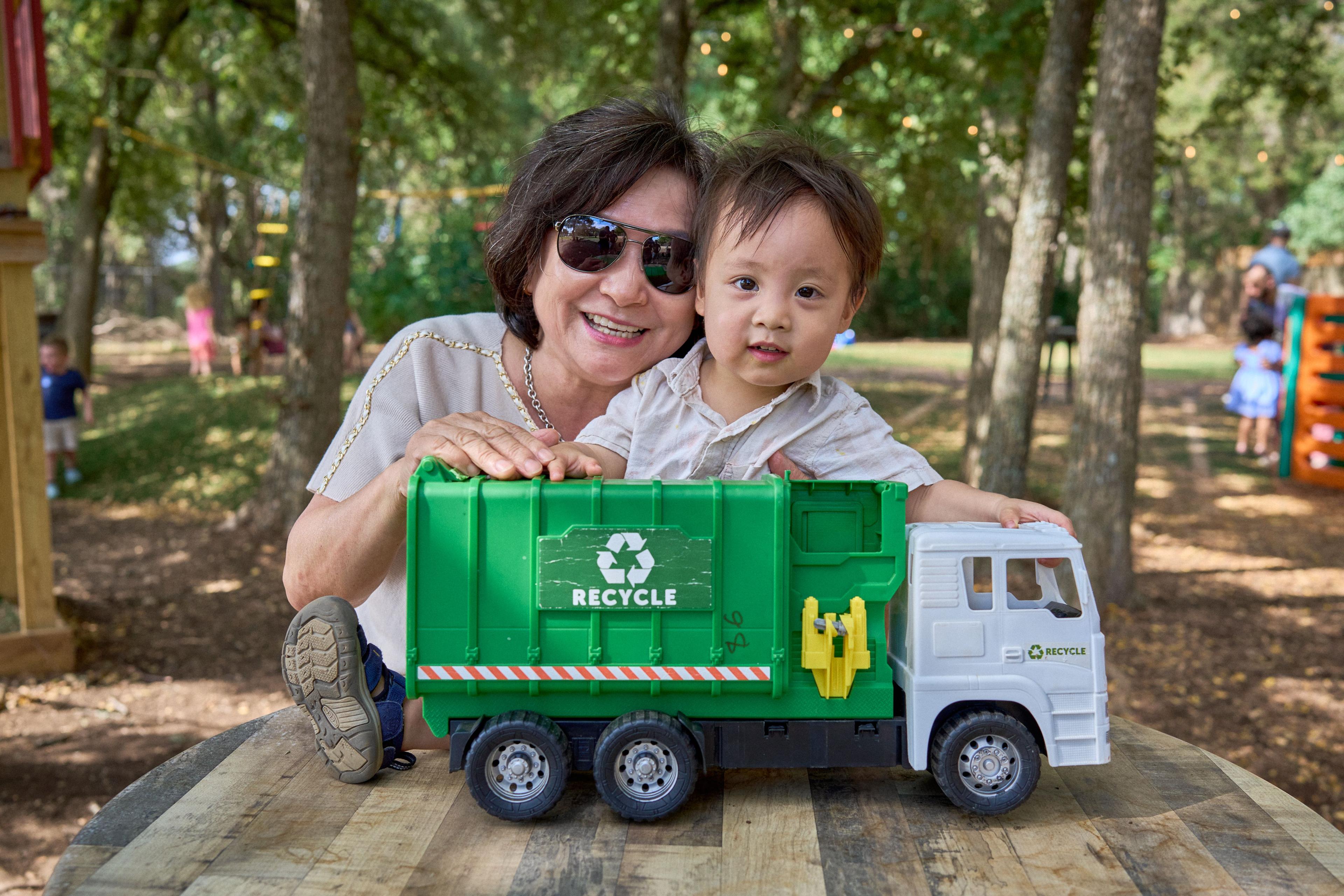 Grandmother in sunglasses holding her grandson, who clutches a green toy recycling truck — Family Parties photography, Austin A grandmother in sunglasses holding her grandson, who clutches a green toy recycling truck.