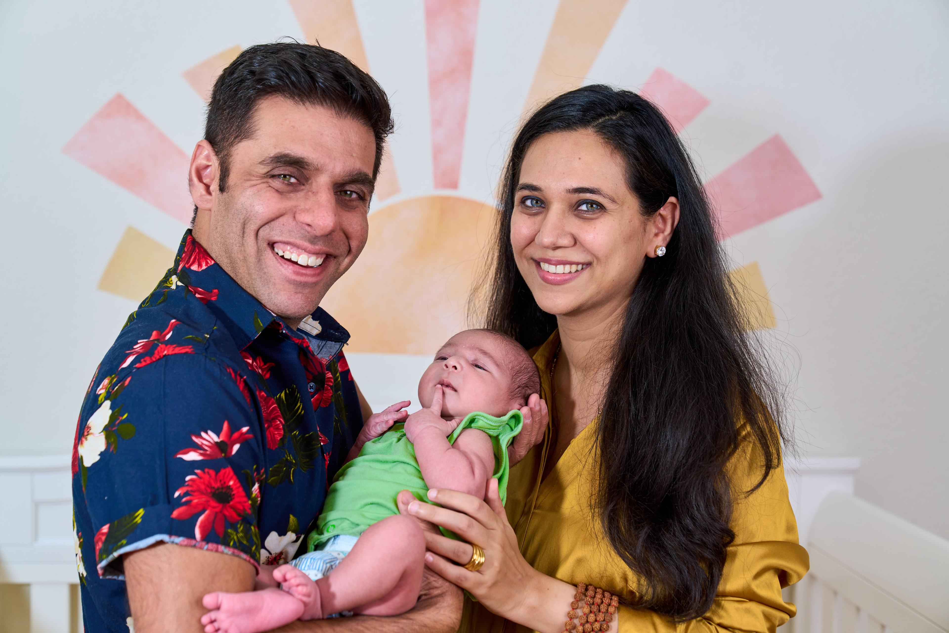 New parents smiling to camera holding their newborn in front of a pastel sunburst mural — Newborn photography, Austin New parents smiling to camera holding their newborn in front of a pastel sunburst mural.