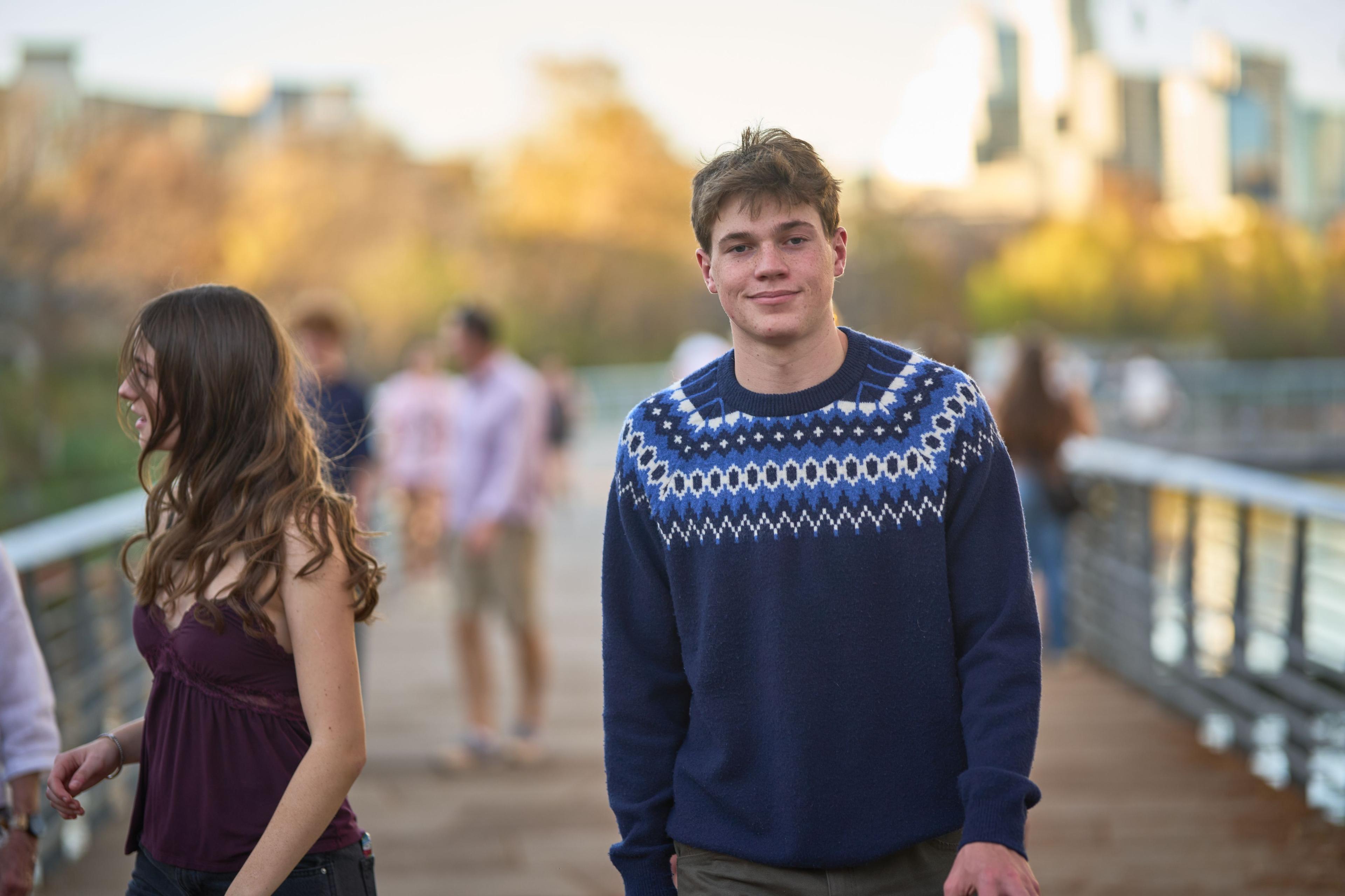 Young man in a patterned navy sweater on the boardwalk path, walkers blurred out behind him — Austin Boardwalk photography, Austin A young man in a patterned navy sweater on the boardwalk path, walkers blurred out behind him in late afternoon light