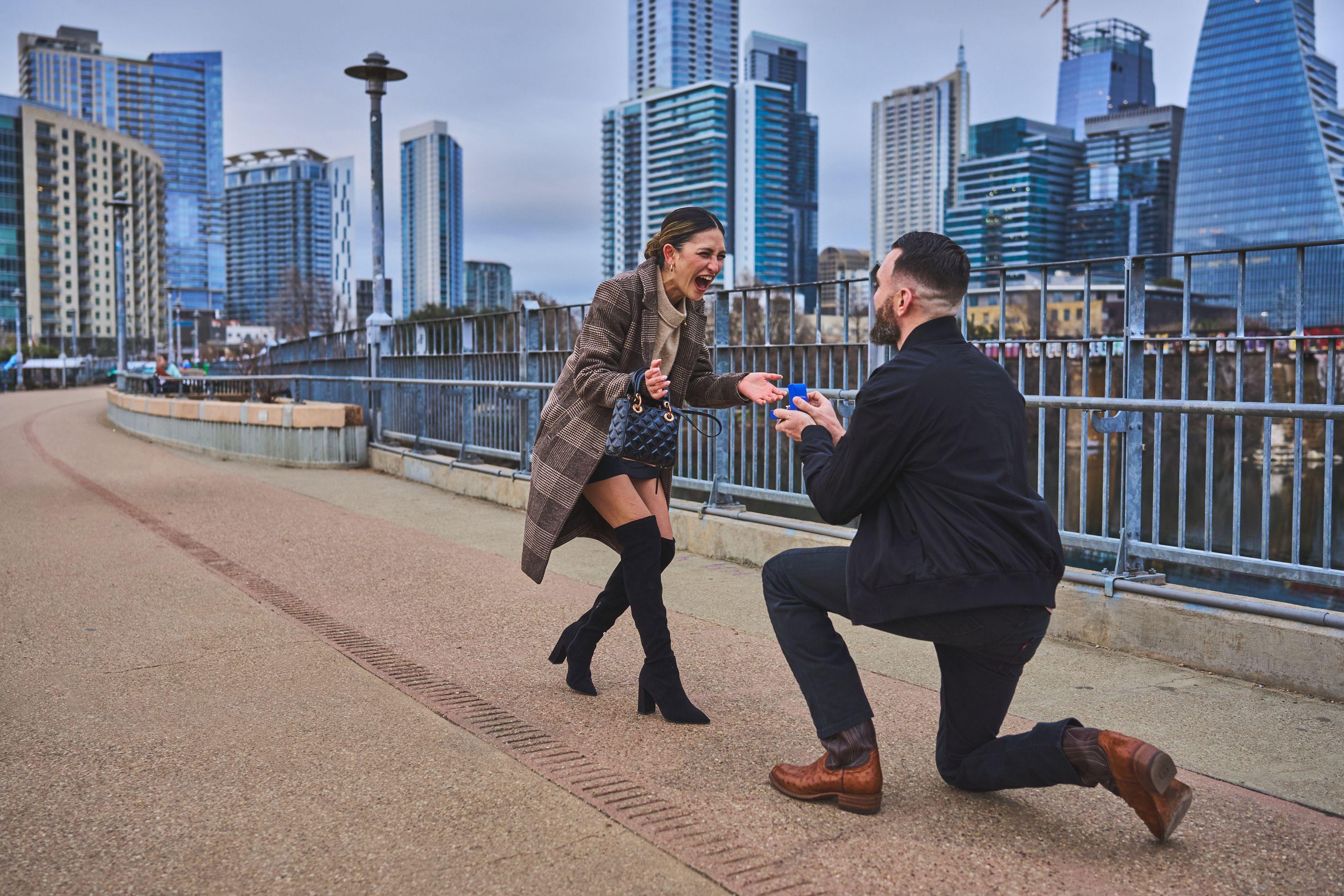 Woman walking up the Pfluger bridge deck toward a man dropping to one knee — Pfluger Pedestrian Bridge photography, Austin A woman walking up the Pfluger bridge deck toward a man dropping to one knee, the downtown Austin skyline behind them