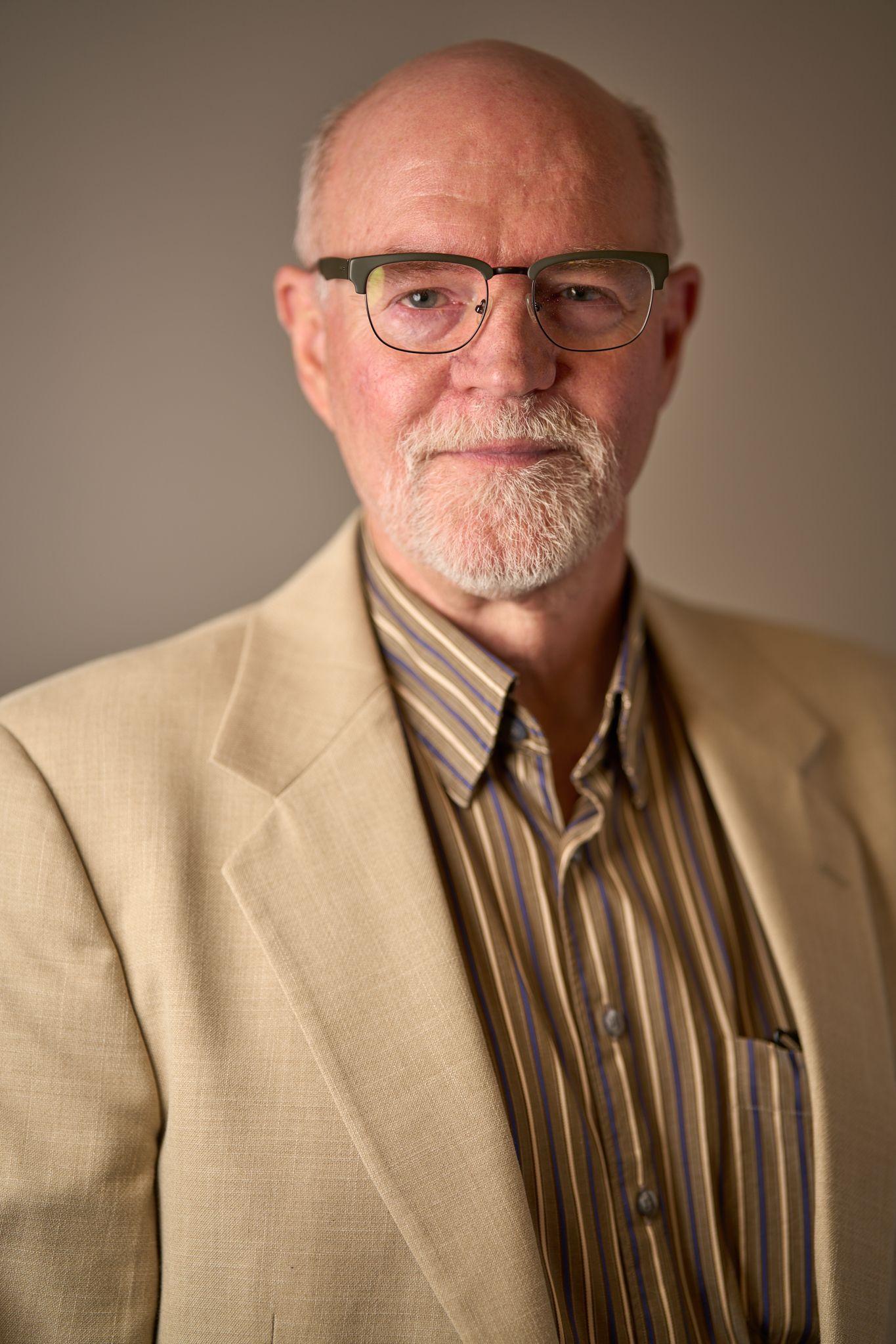 Older man with white hair and glasses in a tan jacket and striped shirt, photographed — Business headshots, Austin An older man with white hair and glasses in a tan jacket and striped shirt, photographed against a dark background