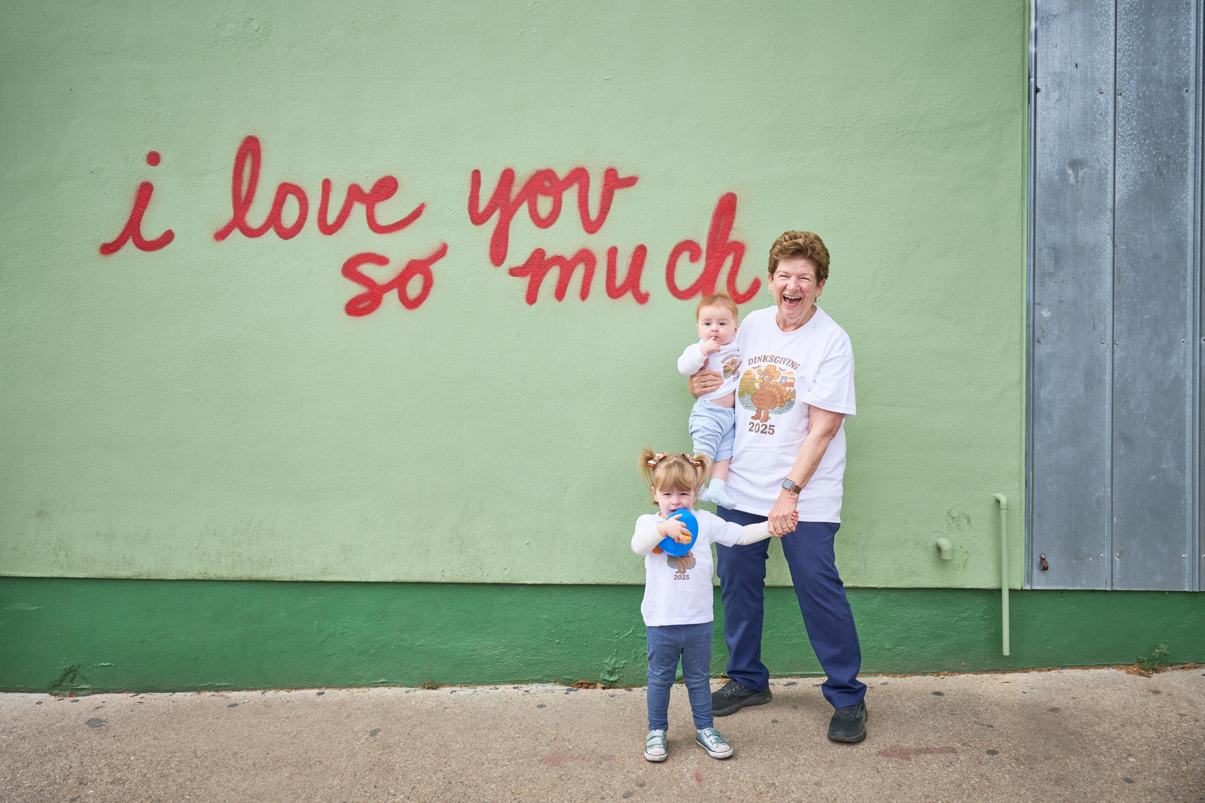 Family with a toddler and baby posed against the green 'love you so much' mural wall — Family Portraits photography, Austin A family with a toddler and baby posed against the green 'love you so much' mural wall.