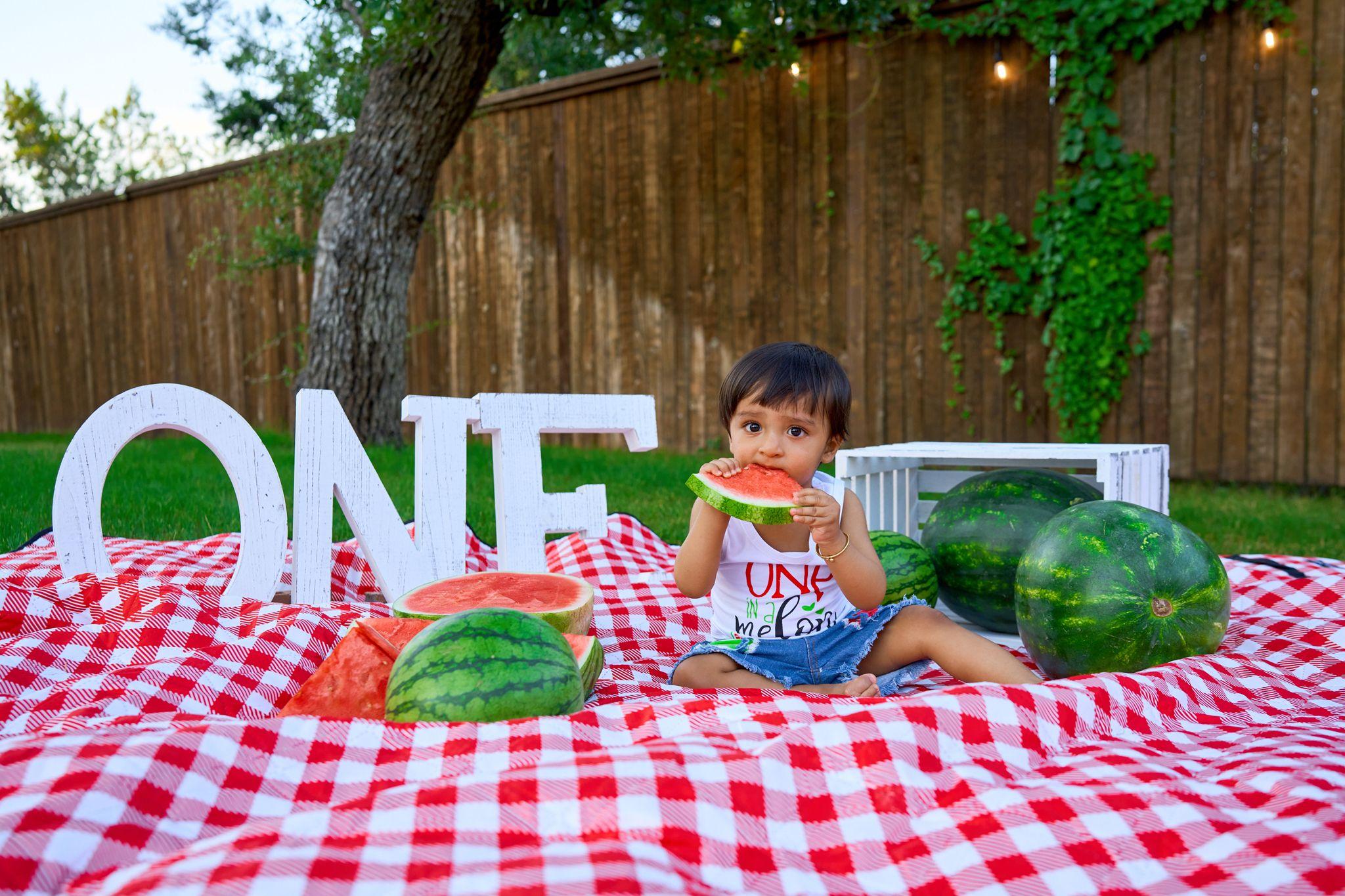 Toddler eating watermelon on a checked picnic blanket beside giant ONE letters — Family Parties photography, Austin A toddler eating watermelon on a checked picnic blanket beside giant ONE letters.