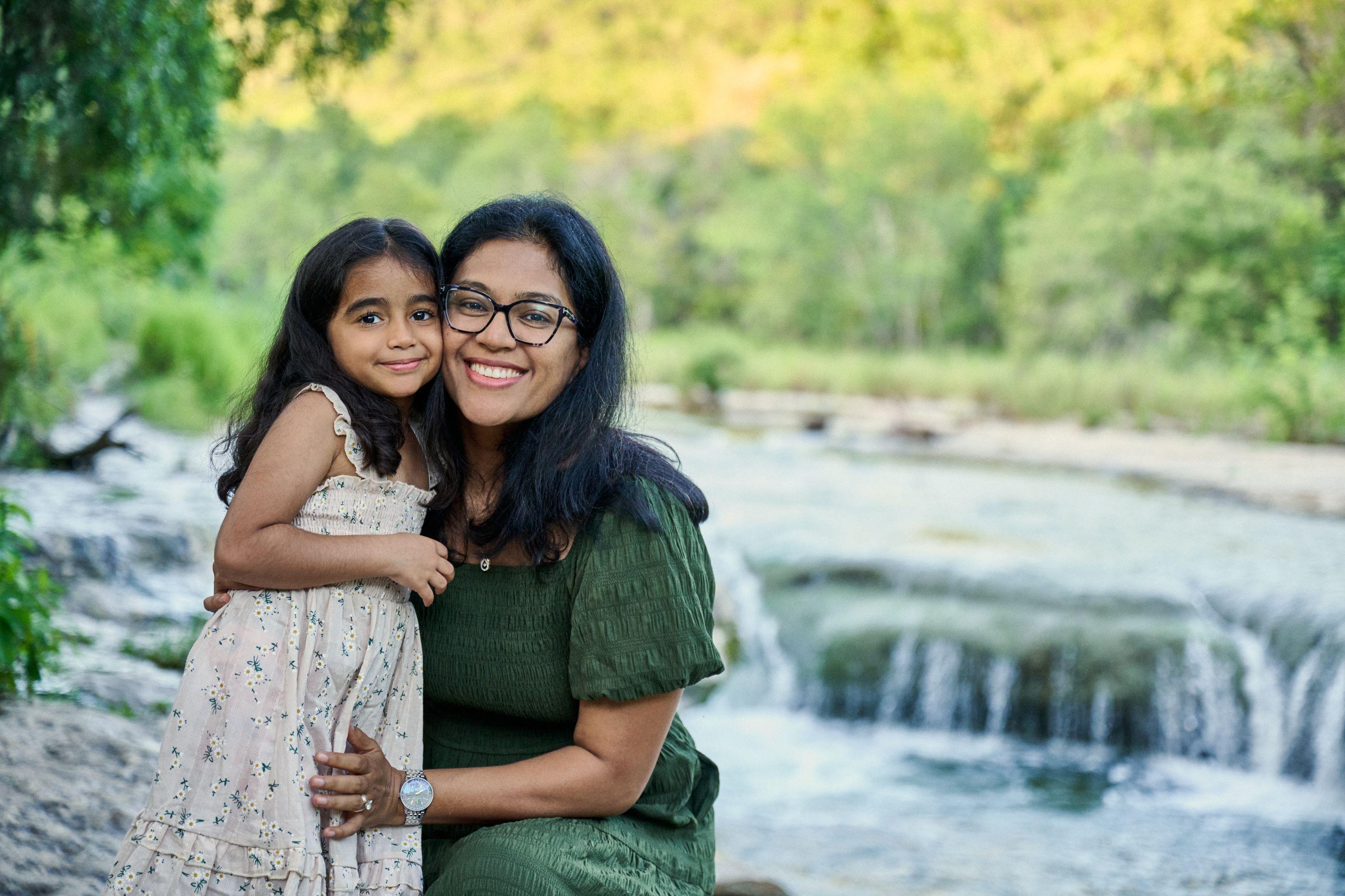 Mother in a green dress holding her daughter with the Bull Creek falls out of focus behind them — Bull Creek District Park photography, Austin A mother in a green dress holding her daughter with the Bull Creek falls out of focus behind them