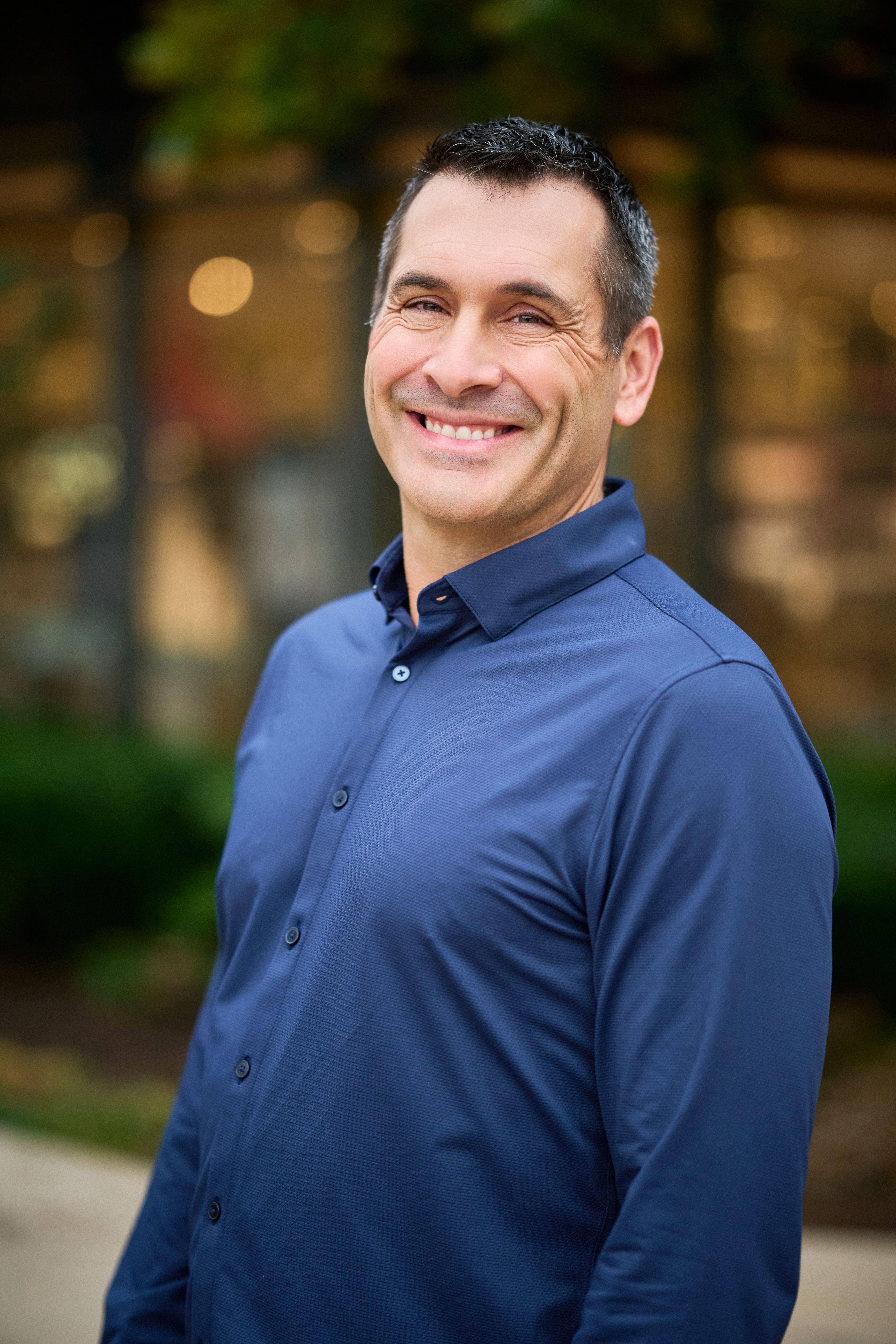 Man in a blue shirt smiling on a Domain street, lit storefronts thrown out of focus behind him — The Domain photography, Austin A man in a blue shirt smiling on a Domain street, lit storefronts thrown out of focus behind him