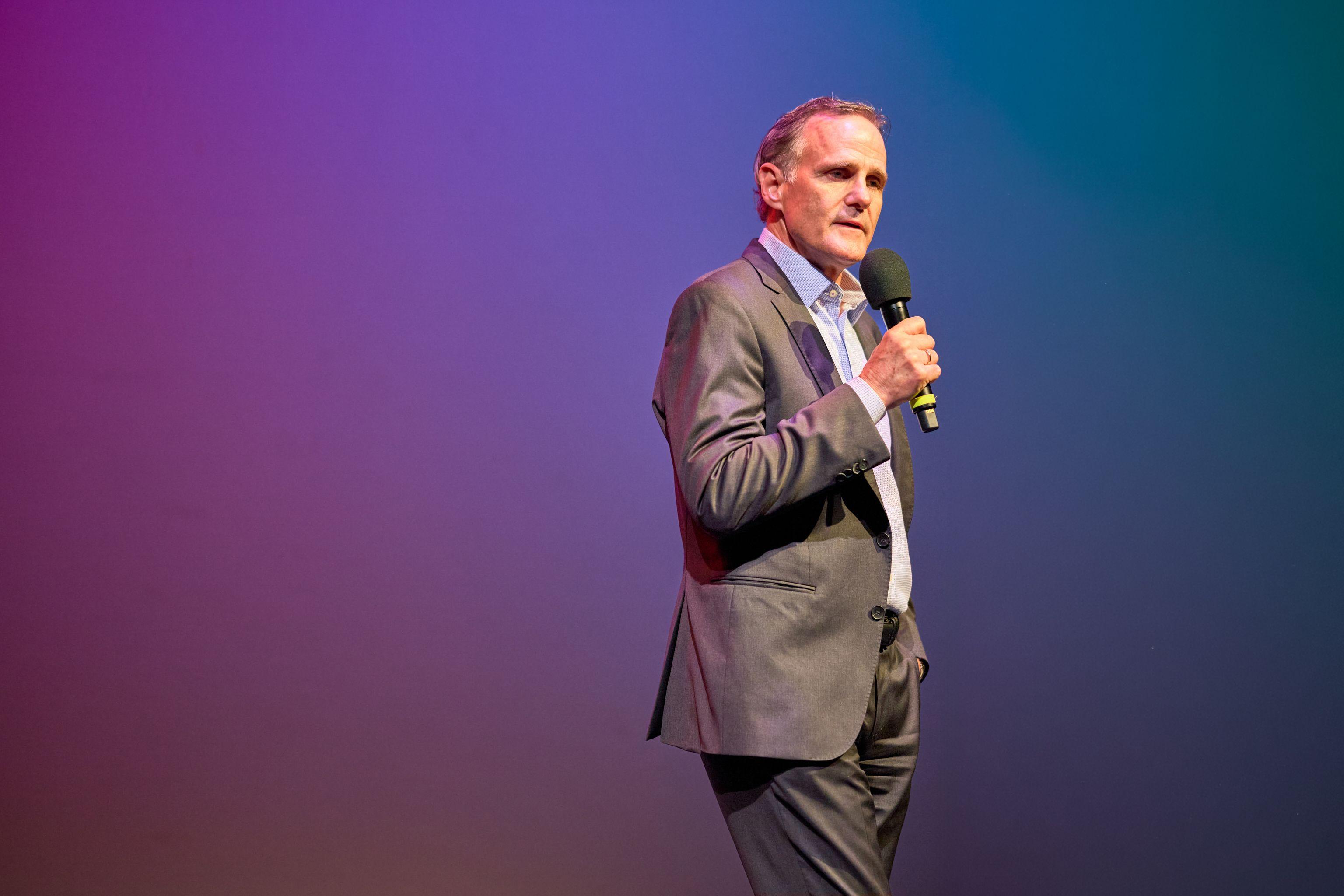 Keynote speaker with a handheld microphone against a magenta and blue gradient screen — Conventions & Tradeshows photography, Austin A keynote speaker with a handheld microphone against a magenta and blue gradient screen.