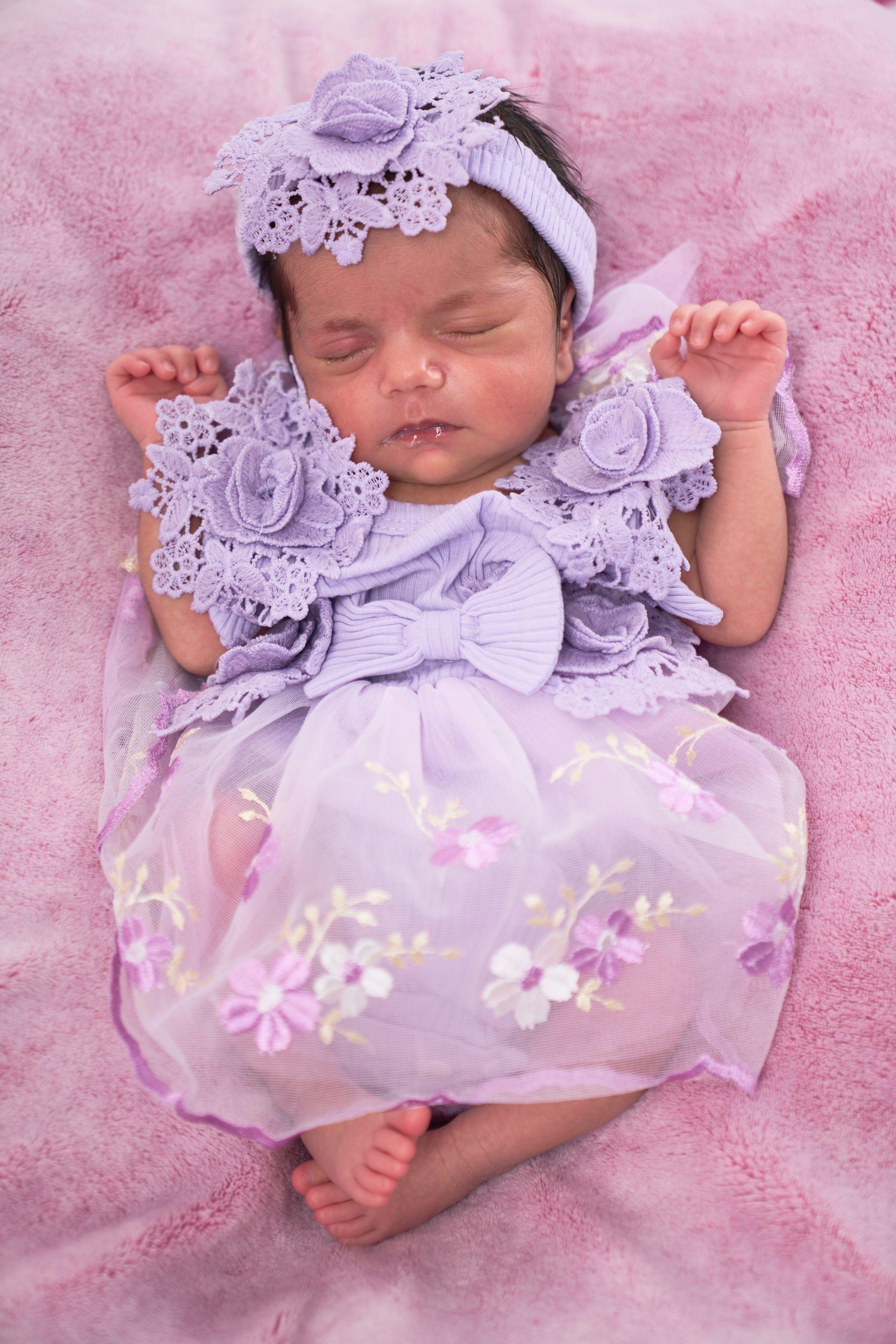 Newborn asleep in a lilac lace dress and floral headband on a pink blanket — Newborn photography, Austin A newborn asleep in a lilac lace dress and floral headband on a pink blanket.
