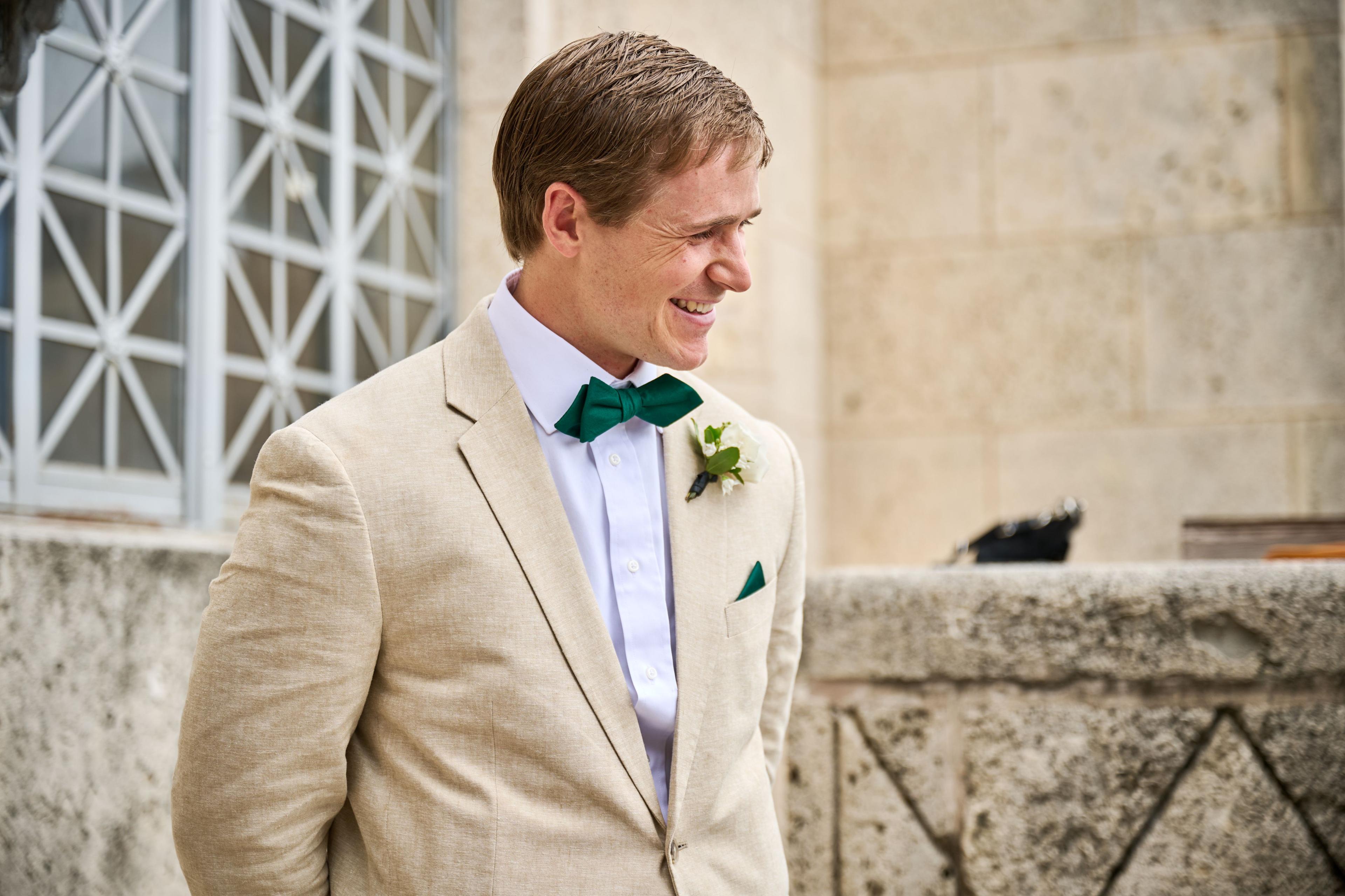 Groom in a cream linen suit smiling against a limestone courthouse wall — Elopements photography, Austin A groom in a cream linen suit smiling against a limestone courthouse wall.