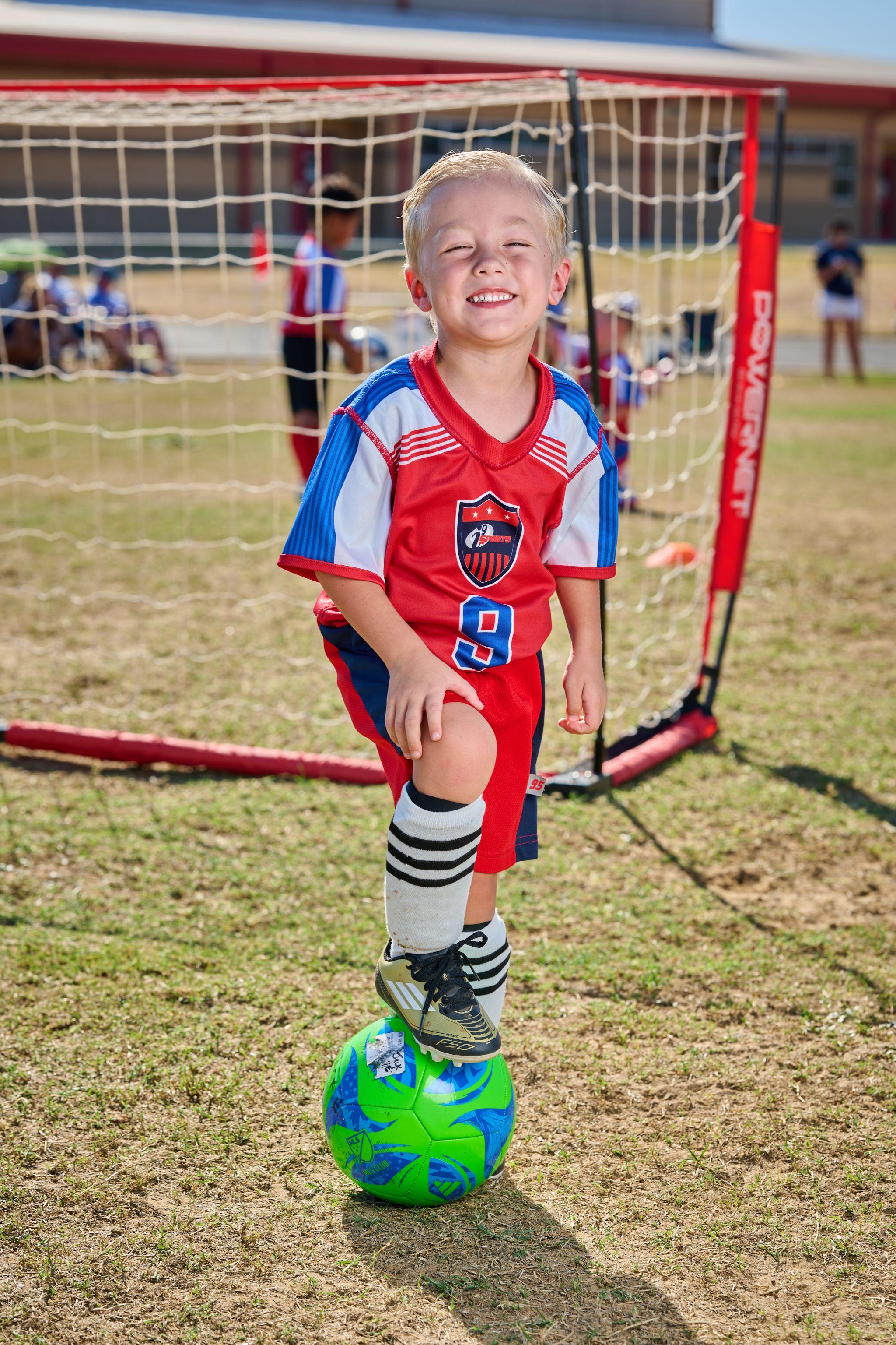 Small child in a red and blue kit standing with one foot on a football, grinning — Sports photography, Austin Small child in a red and blue kit standing with one foot on a football, grinning, in front of a goal