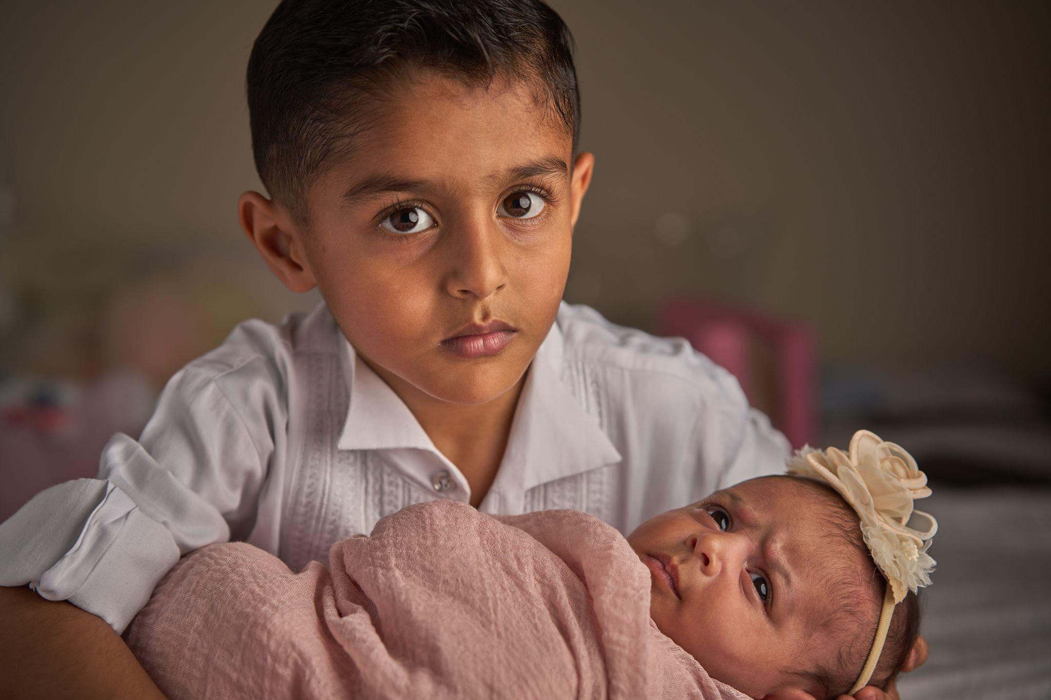 Boy in a white shirt looking to camera while his newborn sibling sleeps below — Newborn photography, Austin A boy in a white shirt looking to camera while his newborn sibling sleeps below.