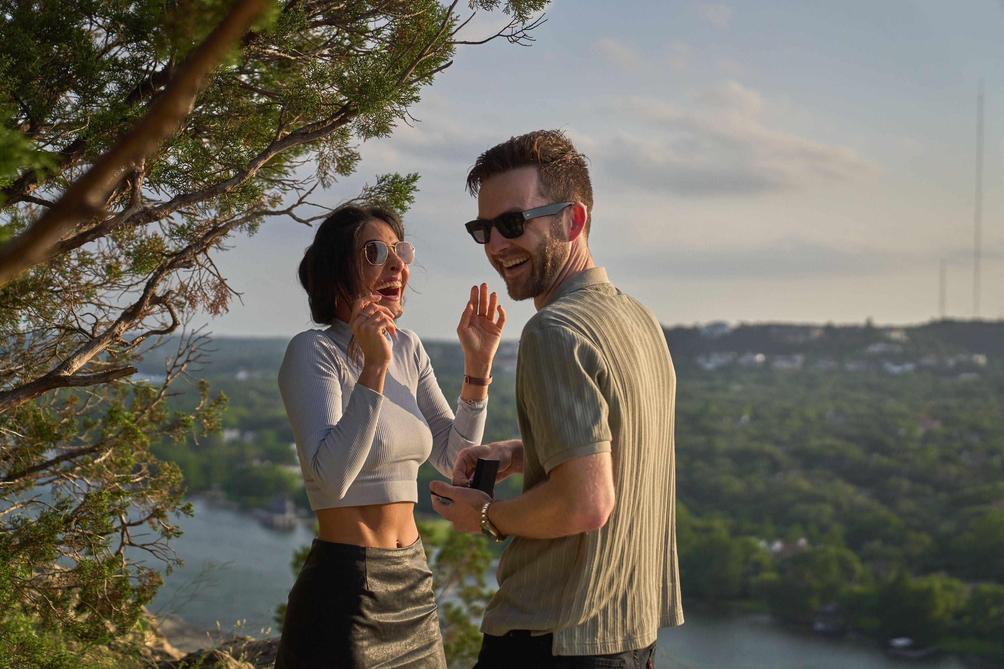 Woman raising her hands to her face in surprise as her partner opens a ring box on the overlook — Engagement & Proposals photography, Austin A woman raising her hands to her face in surprise as her partner opens a ring box on the overlook.
