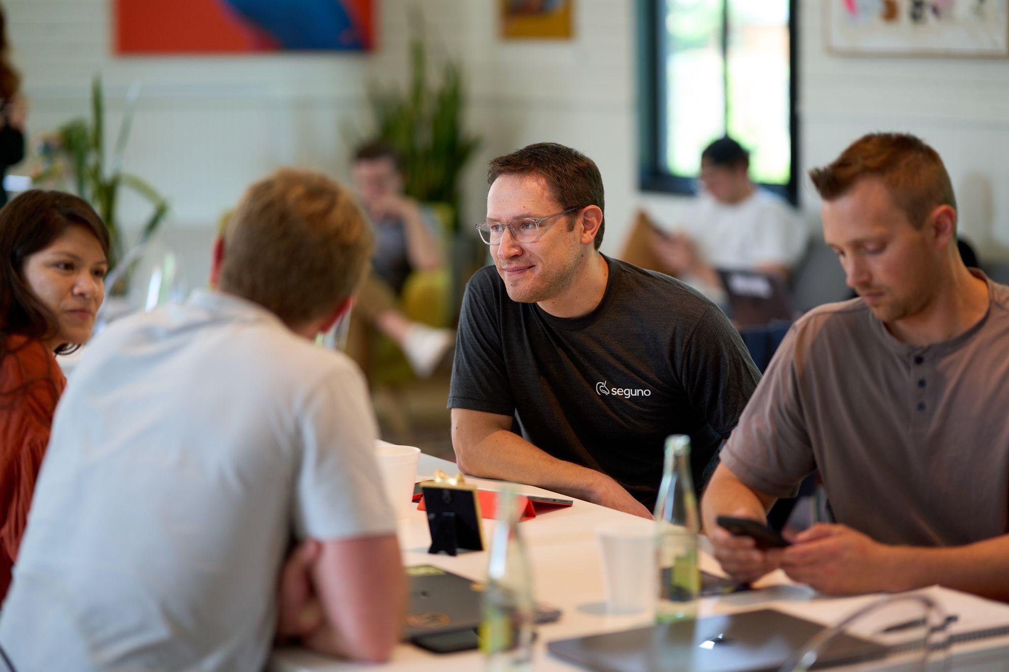 Man in a branded t-shirt laughing during a working session at a long table — Team offsite photography, Austin A man in a branded t-shirt laughing during a working session at a long table