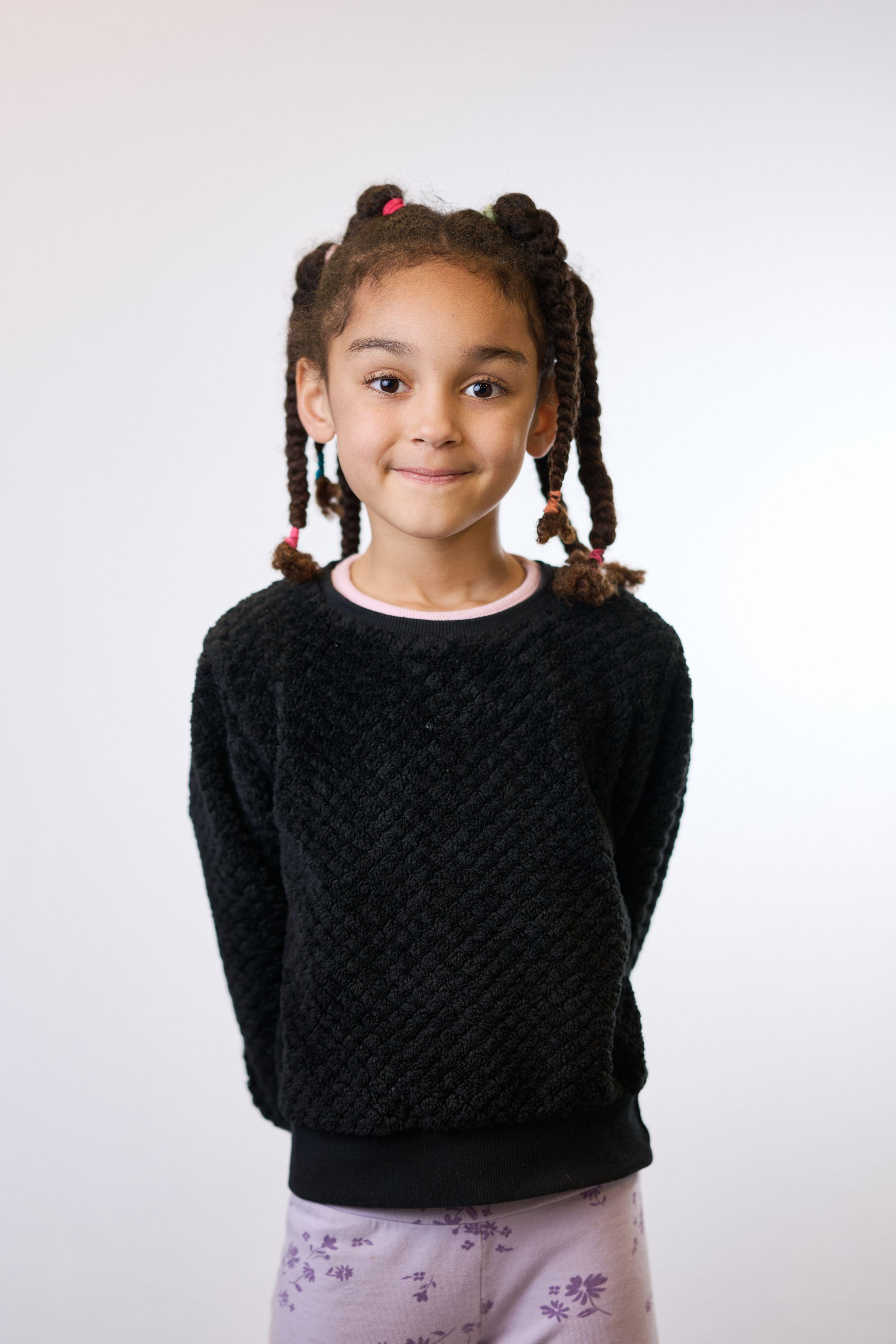 Girl in a black sweater with beaded braids, standing on a plain light backdrop — Mobile photo studio, Austin A girl in a black sweater with beaded braids, standing on a plain light backdrop