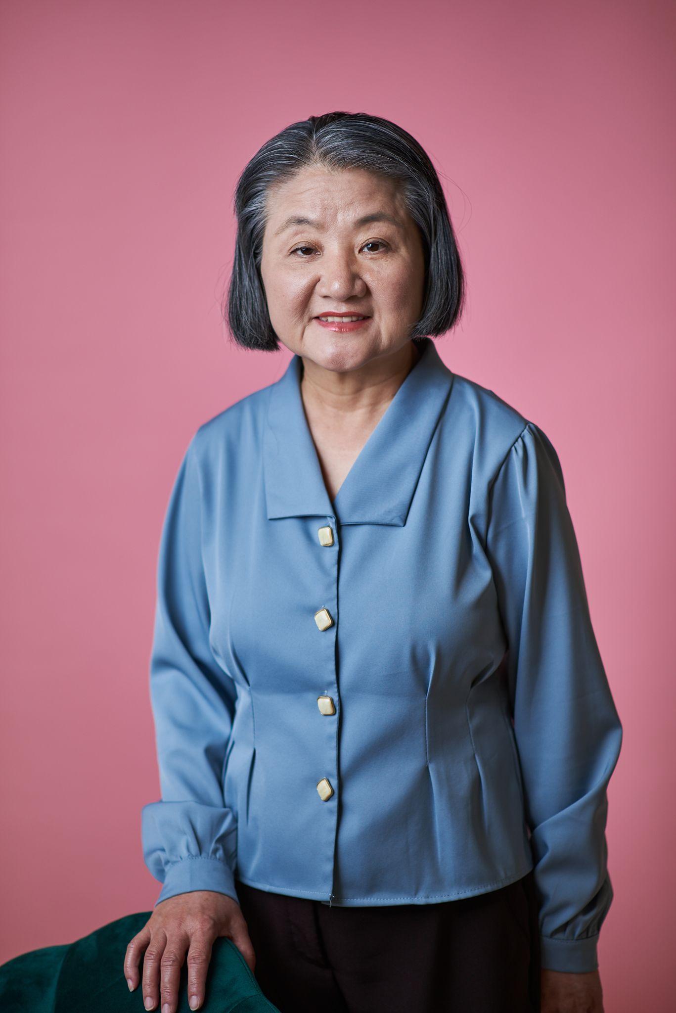 Older woman in a blue blouse smiling against a pink studio backdrop — Headshot photography, Austin An older woman in a blue blouse smiling against a pink studio backdrop.