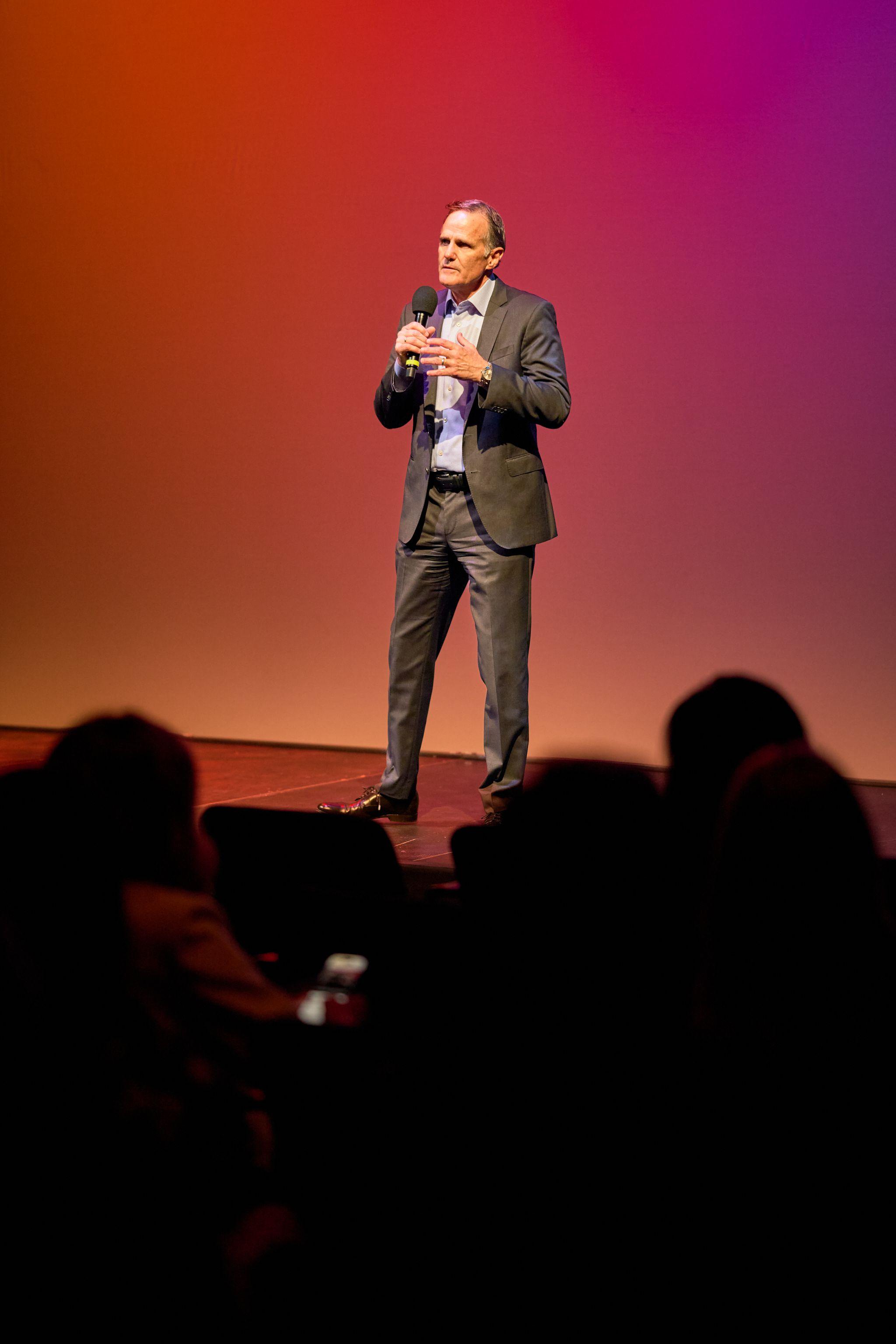 Speaker on a red-lit stage addressing a silhouetted audience — Conventions & Tradeshows photography, Austin A speaker on a red-lit stage addressing a silhouetted audience.