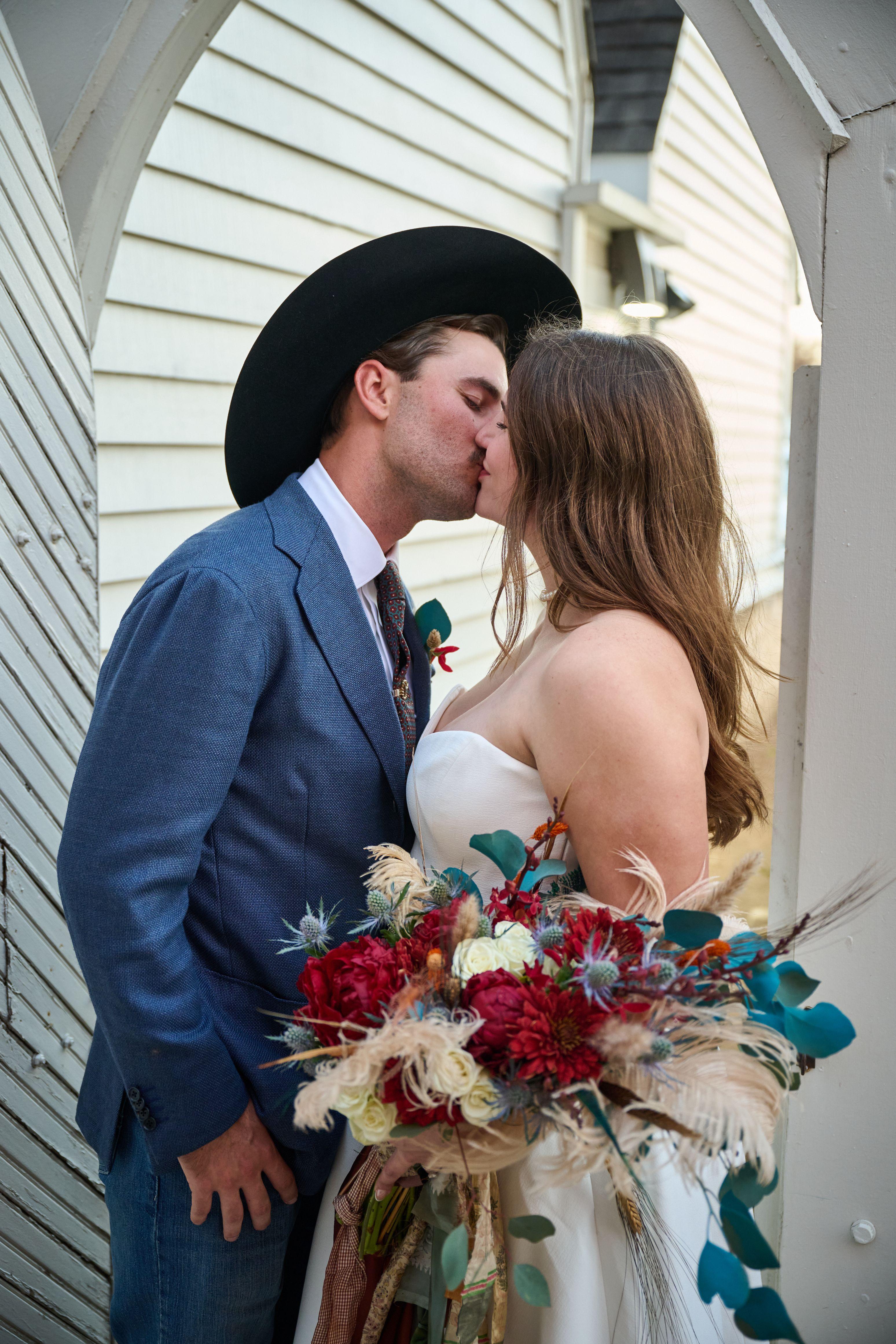 Groom in a black cowboy hat kissing his bride, who holds a deep red and teal bouquet — Weddings photography, Austin A groom in a black cowboy hat kissing his bride, who holds a deep red and teal bouquet, beside a white clapboard wall.