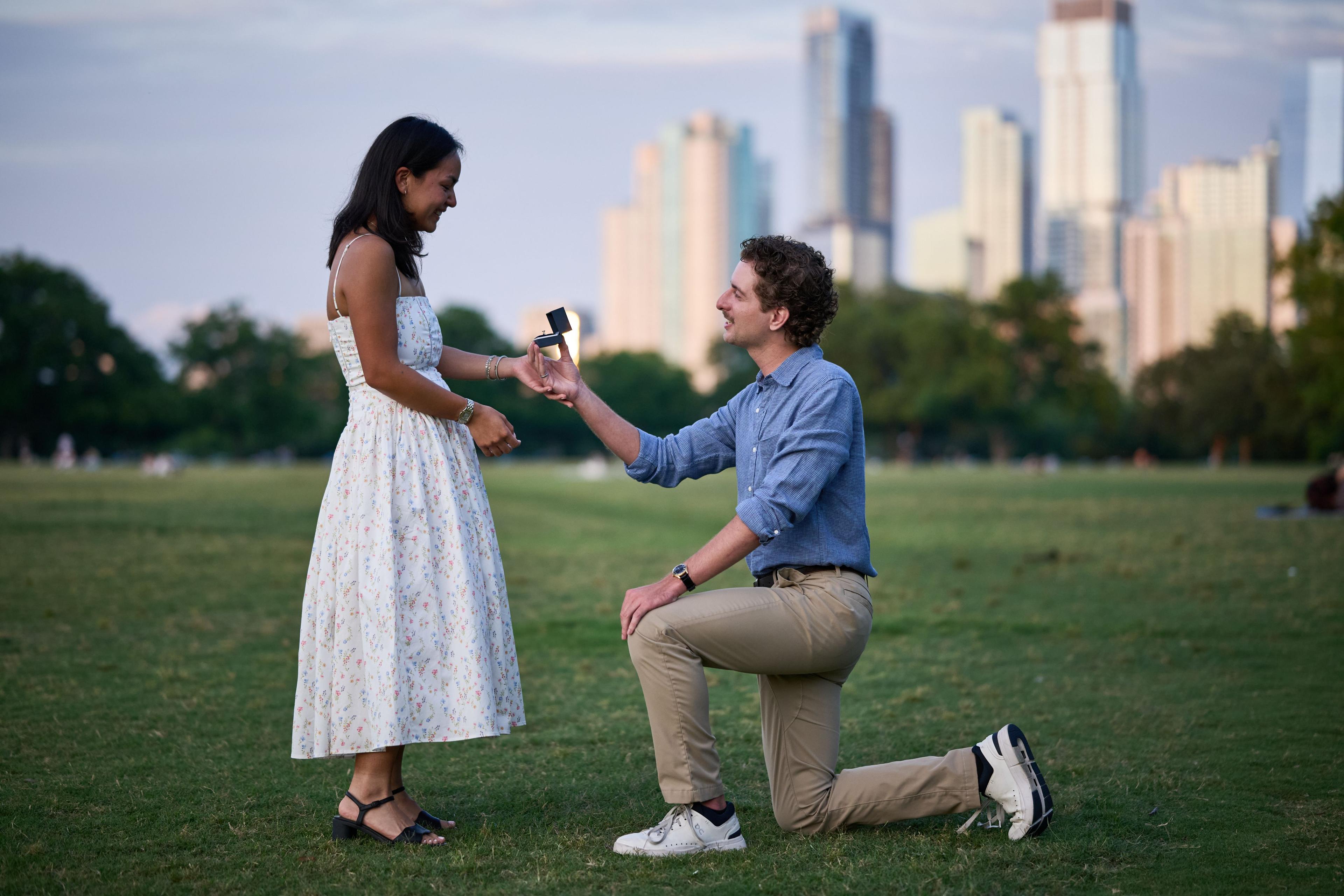 Man on one knee proposing to a woman in a white dress on the Zilker Park lawn, their dog — Zilker Park photography, Austin A man on one knee proposing to a woman in a white dress on the Zilker Park lawn, their dog beside them and the downtown skyline across the water