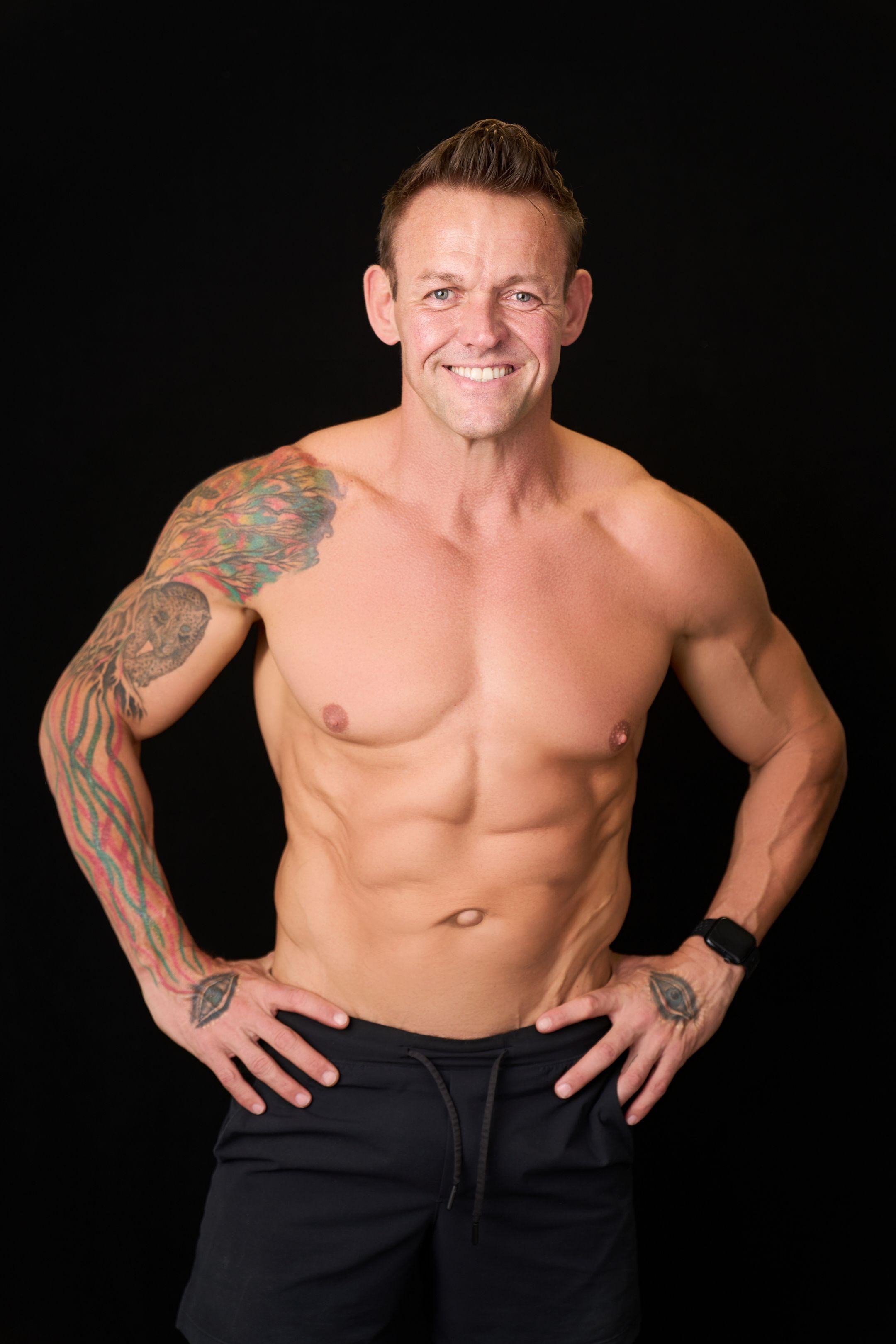 A muscular tattooed man photographed shirtless with hands on hips against a black studio background