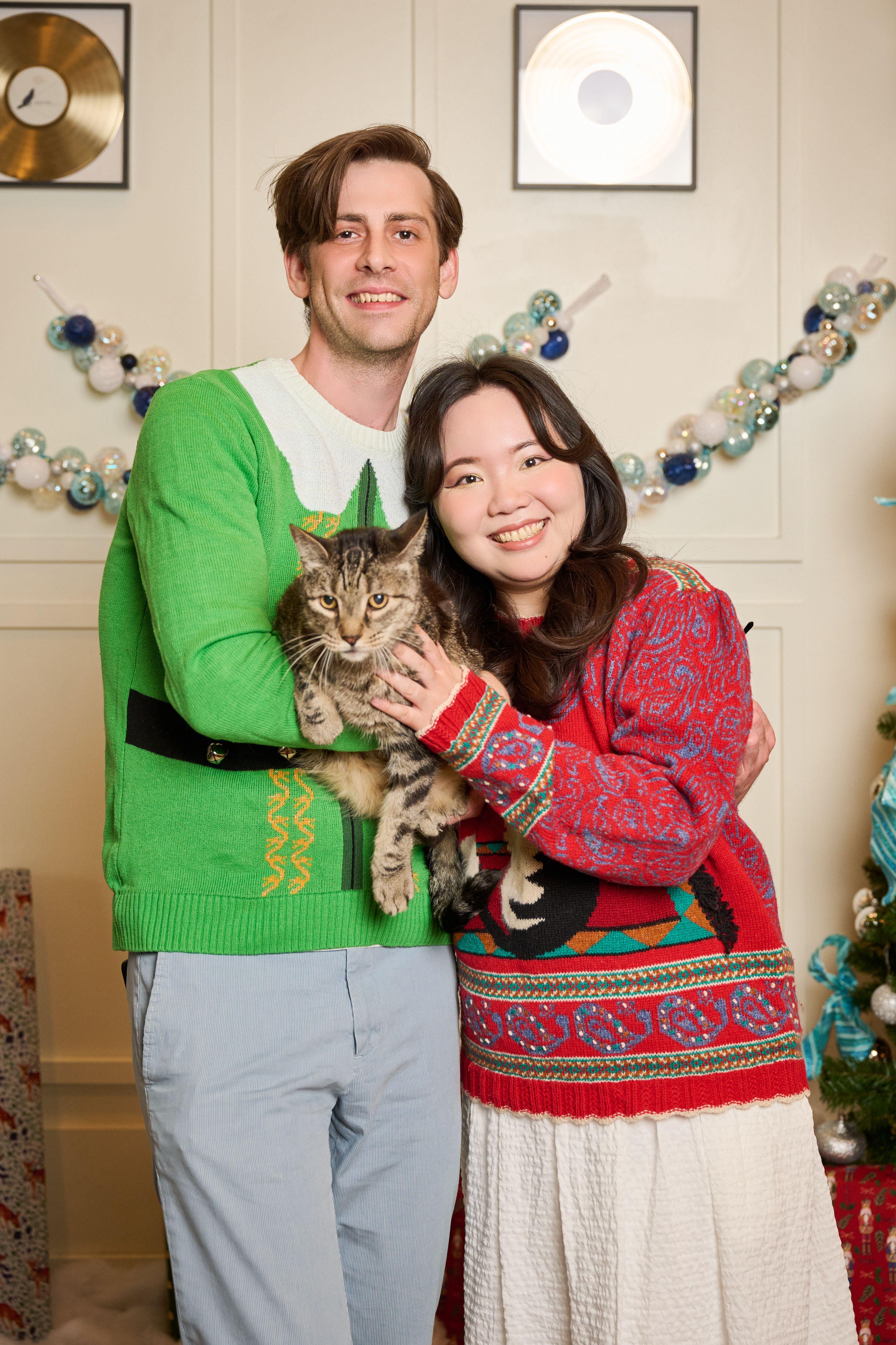 Couple in bright knitted holiday sweaters holding a tabby cat in front of a decorated wall — Mobile photo studio, Austin A couple in bright knitted holiday sweaters holding a tabby cat in front of a decorated wall