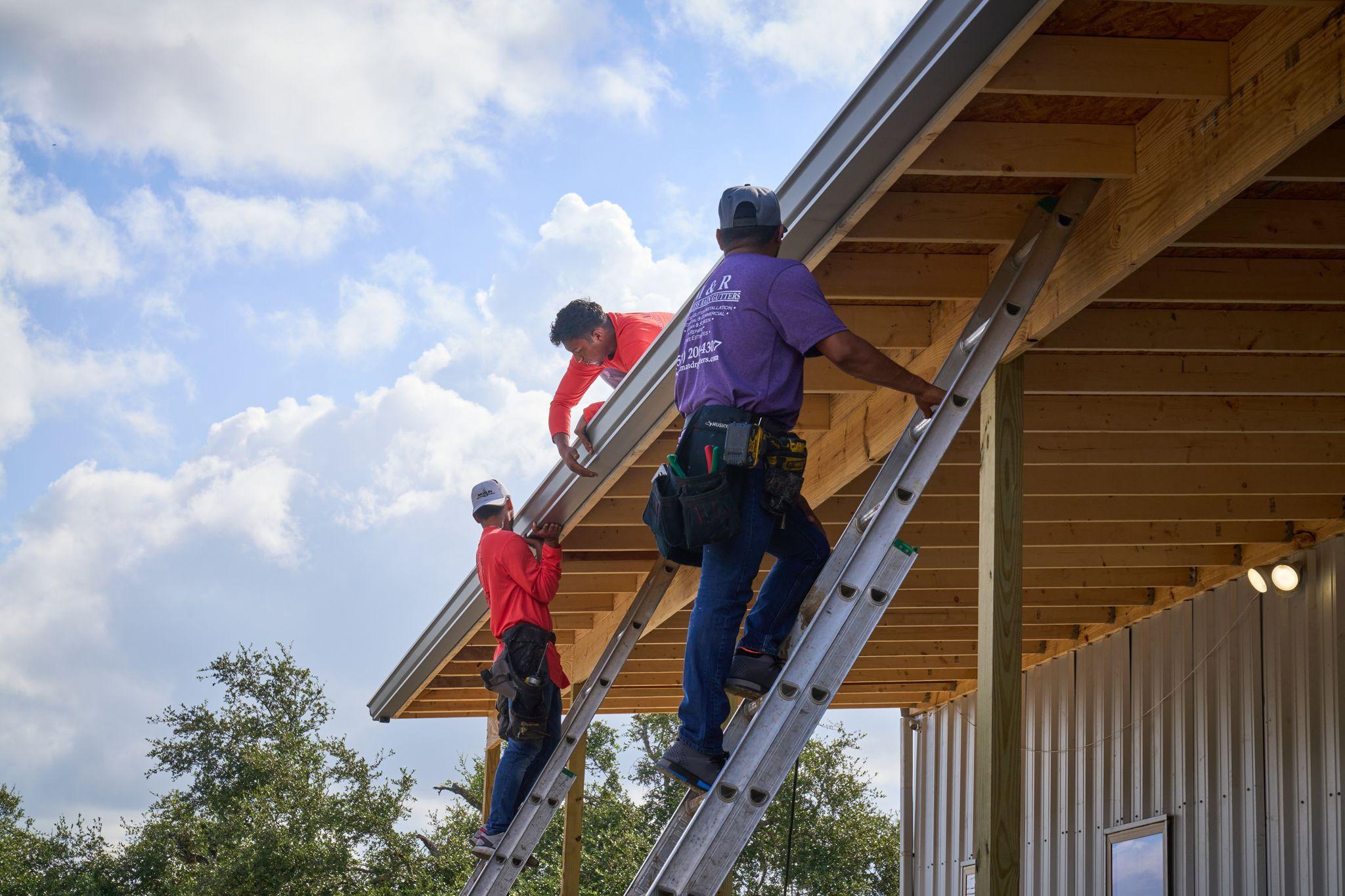 Two roofers on ladders fitting timber and metal trusses on a new roof against a blue sky — Industrial photography, Austin Two roofers on ladders fitting timber and metal trusses on a new roof against a blue sky