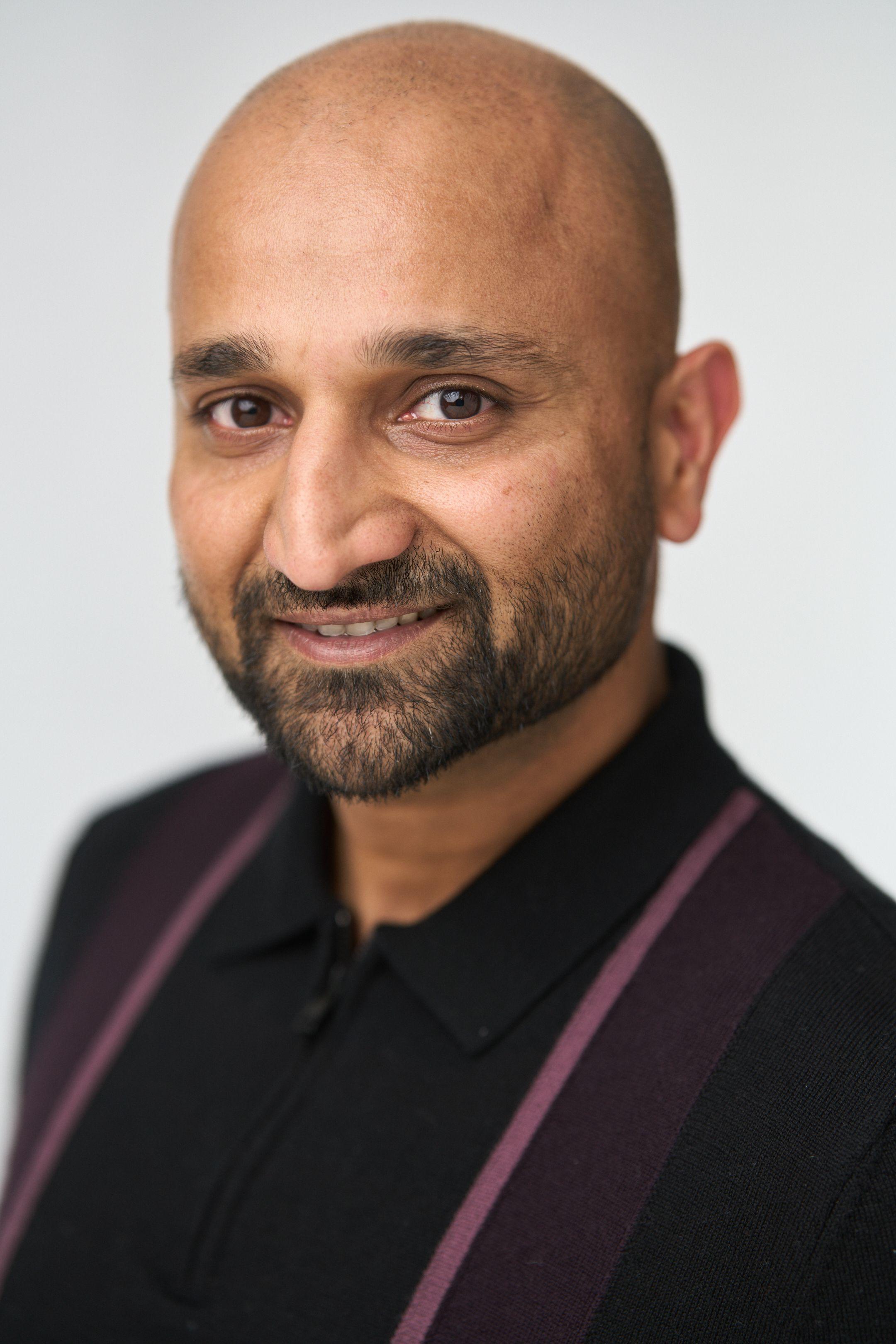 Close-cropped headshot of a bald man with a beard in a dark polo and striped jacket — Business headshots, Austin A close-cropped headshot of a bald man with a beard in a dark polo and striped jacket against a pale gray background