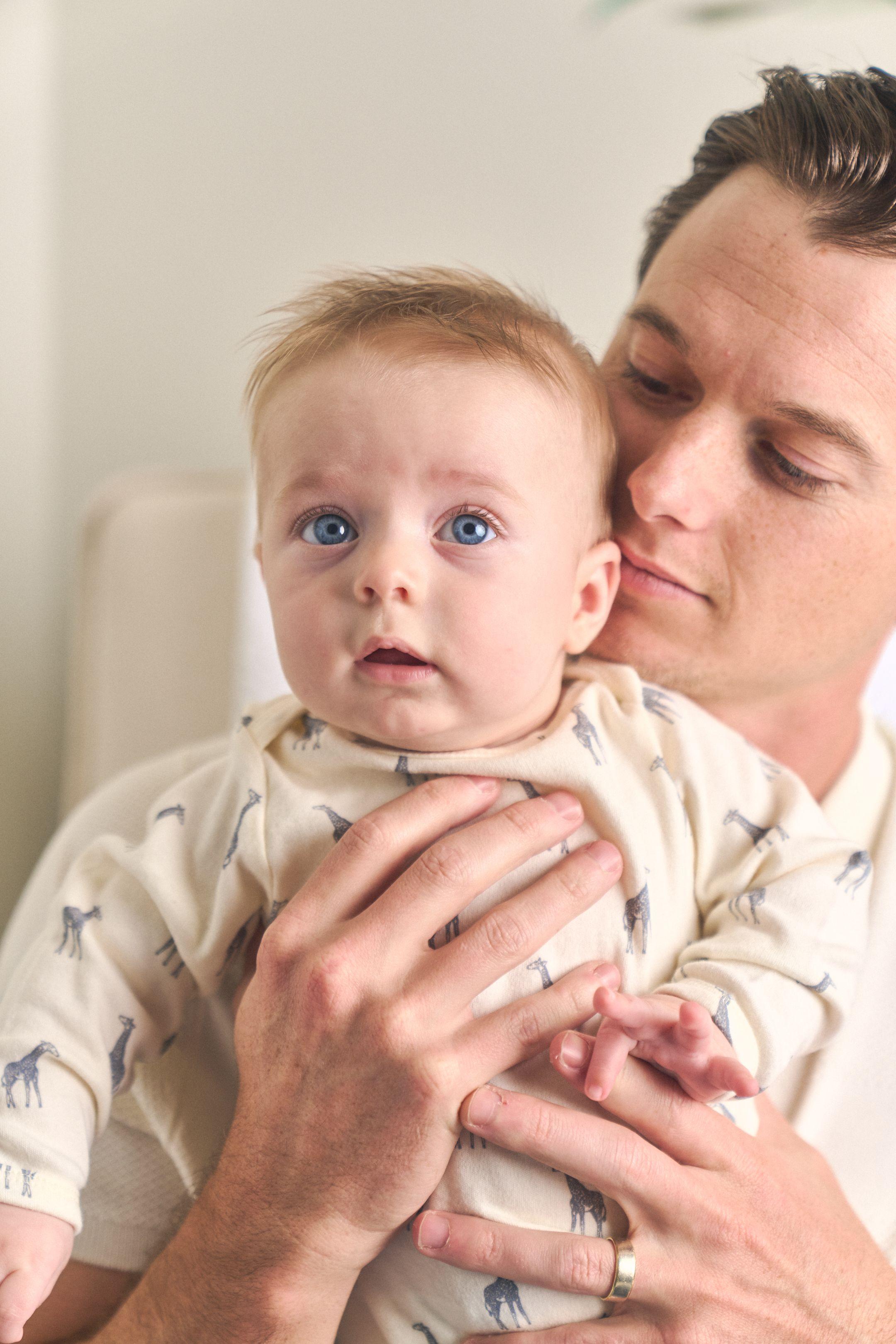 Baby in a giraffe-print sleepsuit held against his father's chest, looking to camera — Newborn photography, Austin A baby in a giraffe-print sleepsuit held against his father's chest, looking to camera.