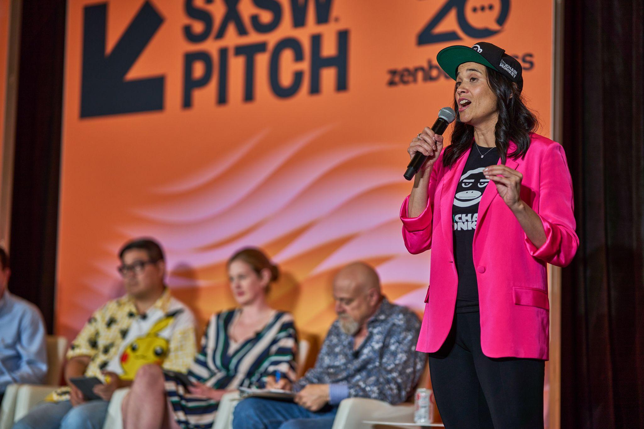 Host in a pink blazer speaking into a microphone in front of the orange SXSW Pitch backdrop — Conventions & Tradeshows photography, Austin A host in a pink blazer speaking into a microphone in front of the orange SXSW Pitch backdrop.