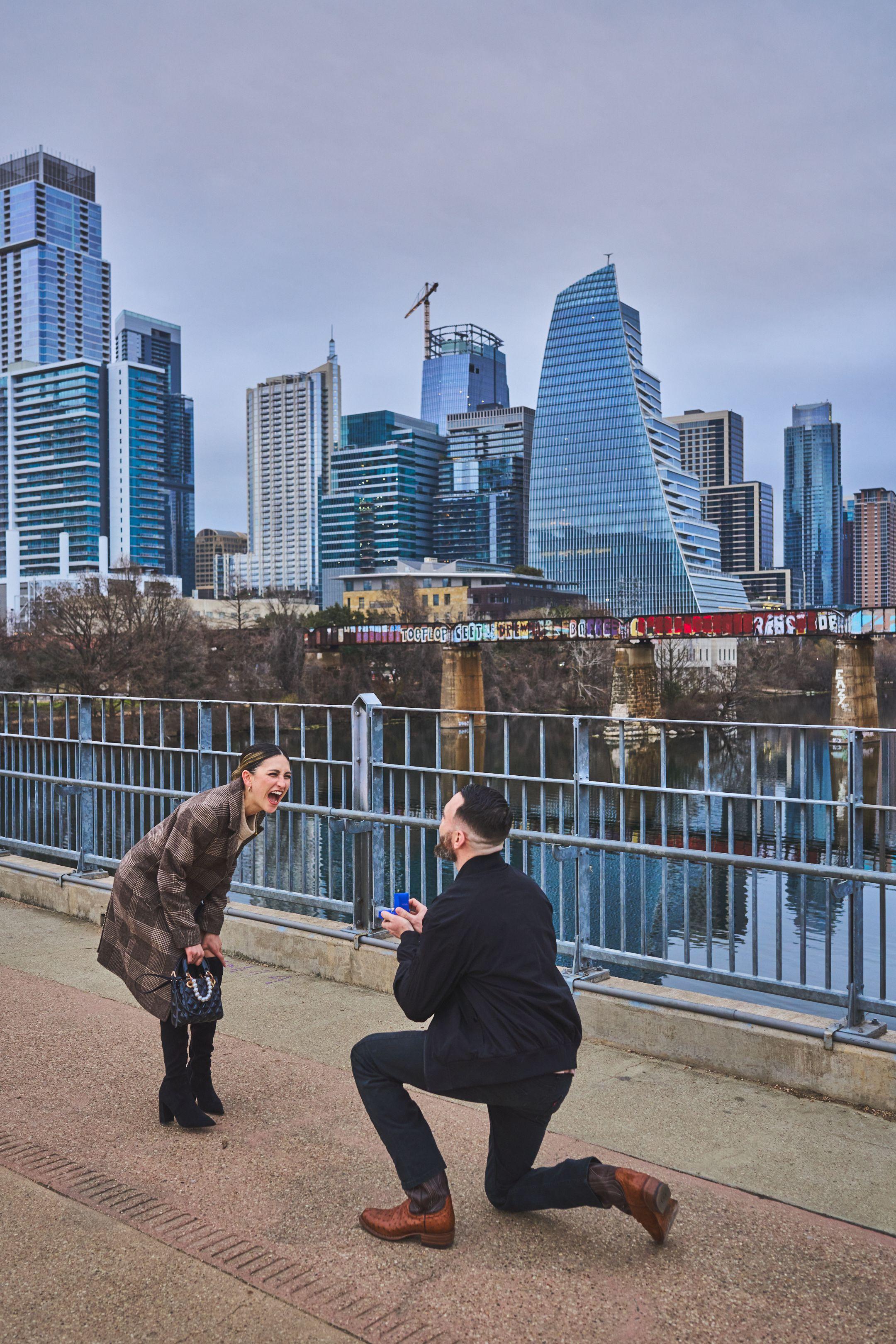 Man on one knee holding a ring box on the Pfluger bridge, the woman's hands at her face — Pfluger Pedestrian Bridge photography, Austin A man on one knee holding a ring box on the Pfluger bridge, the woman's hands at her face and the skyline across the water behind