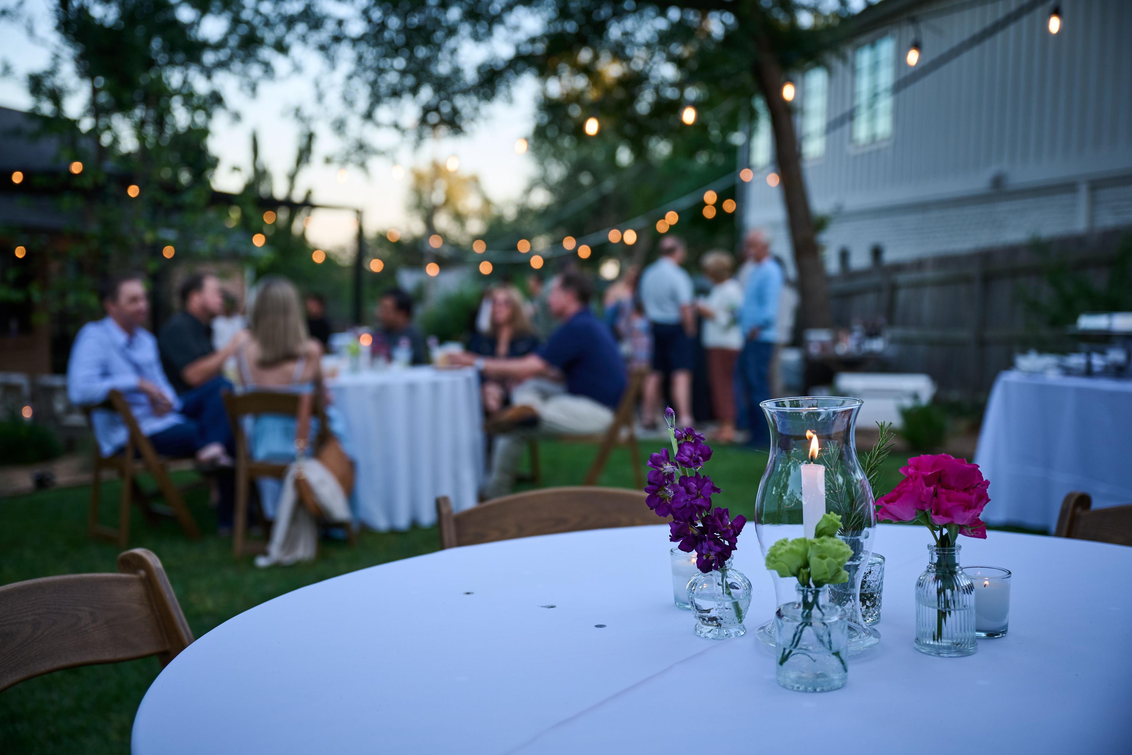 Guests at round tables on a lawn under string lights at dusk, candles and flowers on the linen — Formal Celebrations photography, Austin Guests at round tables on a lawn under string lights at dusk, candles and flowers on the linen.