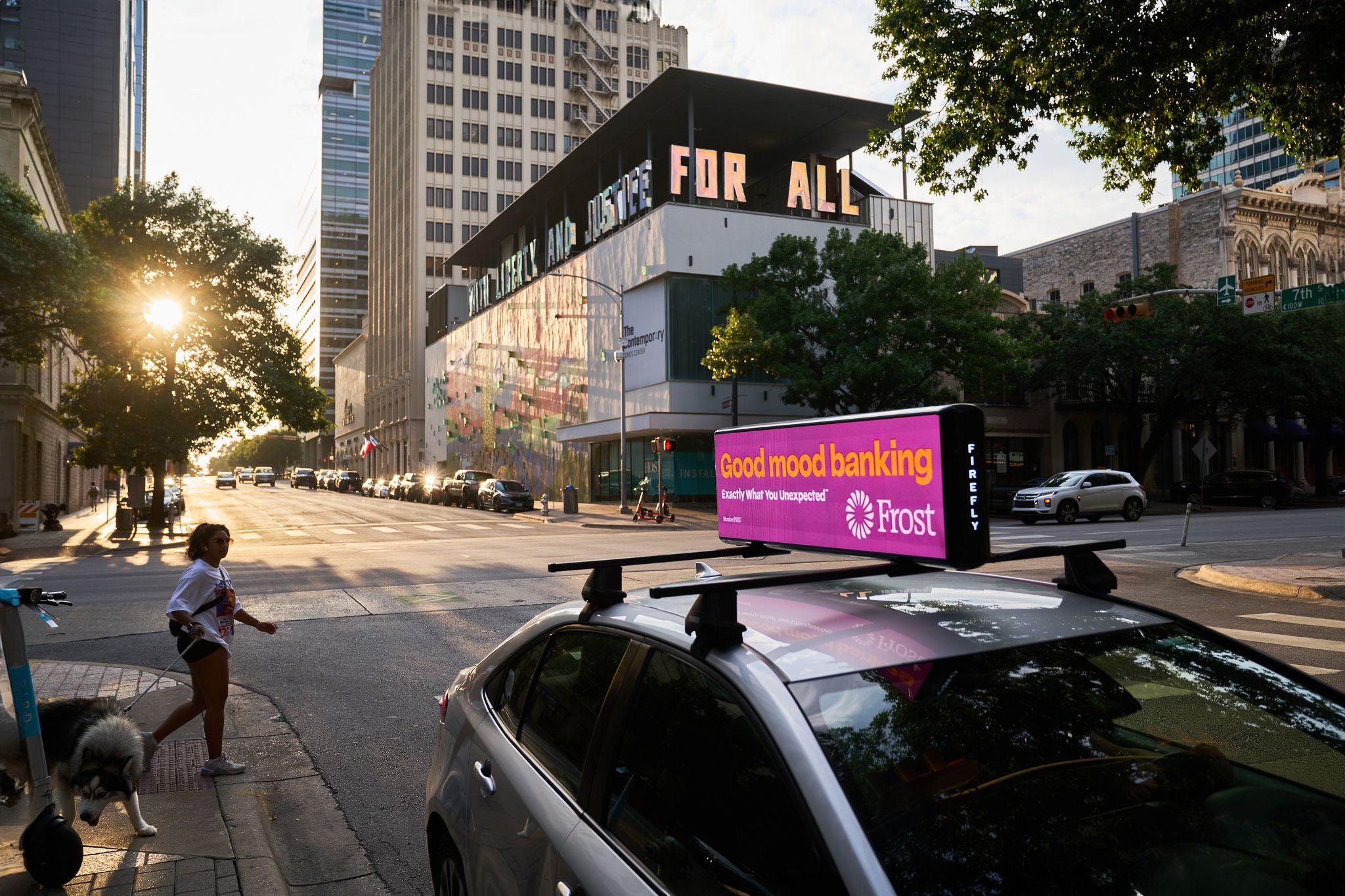 Car-top digital screen running a magenta Frost Bank 'Good mood banking' ad on a downtown street — Downtown Austin photography, Austin A car-top digital screen running a magenta Frost Bank 'Good mood banking' ad on a downtown street, below a building lettered FOR ALL.
