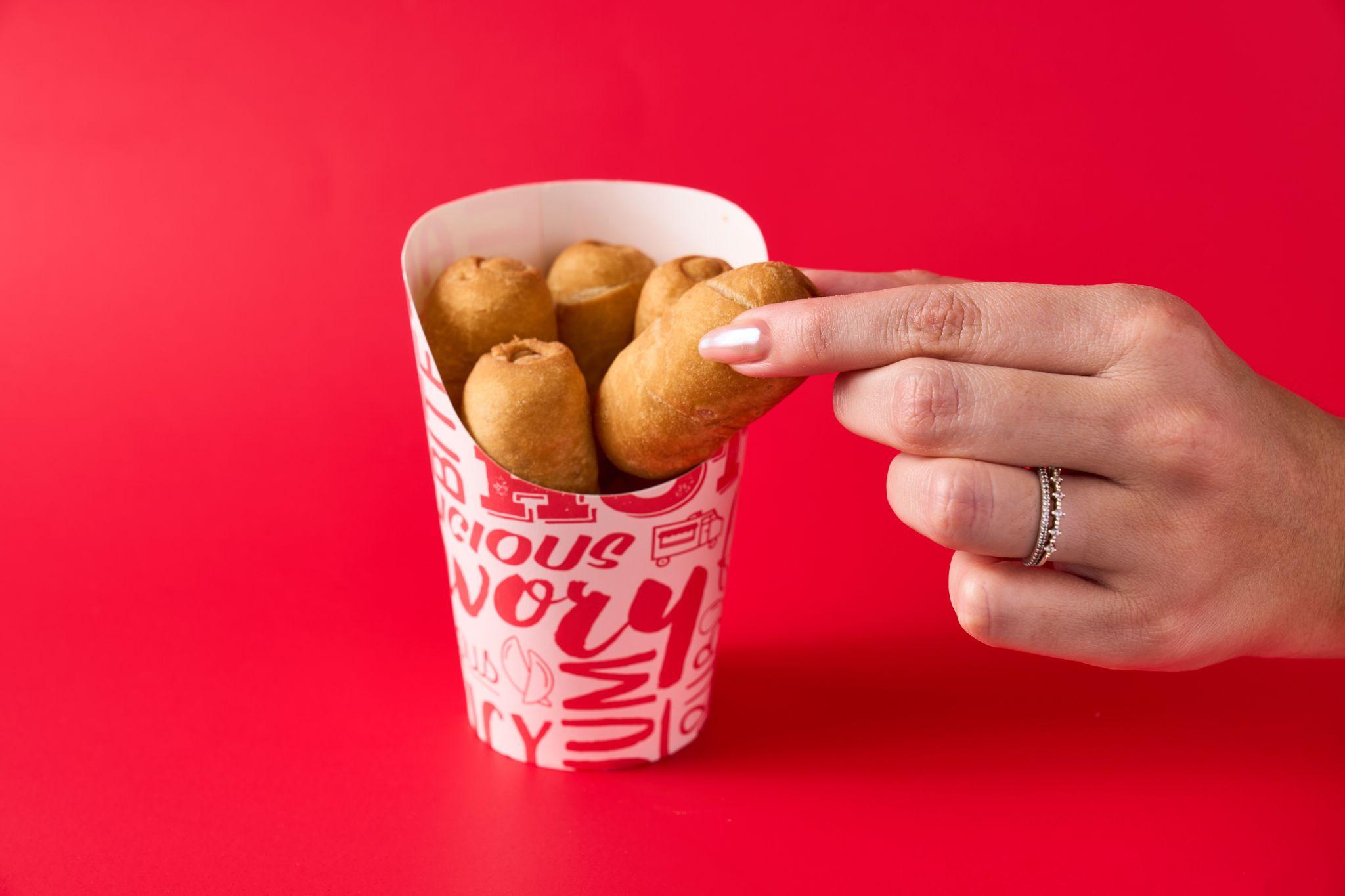 Hand reaching into a branded cup of corn dog bites against a red background — Product photography, Austin A hand reaching into a branded cup of corn dog bites against a red background.