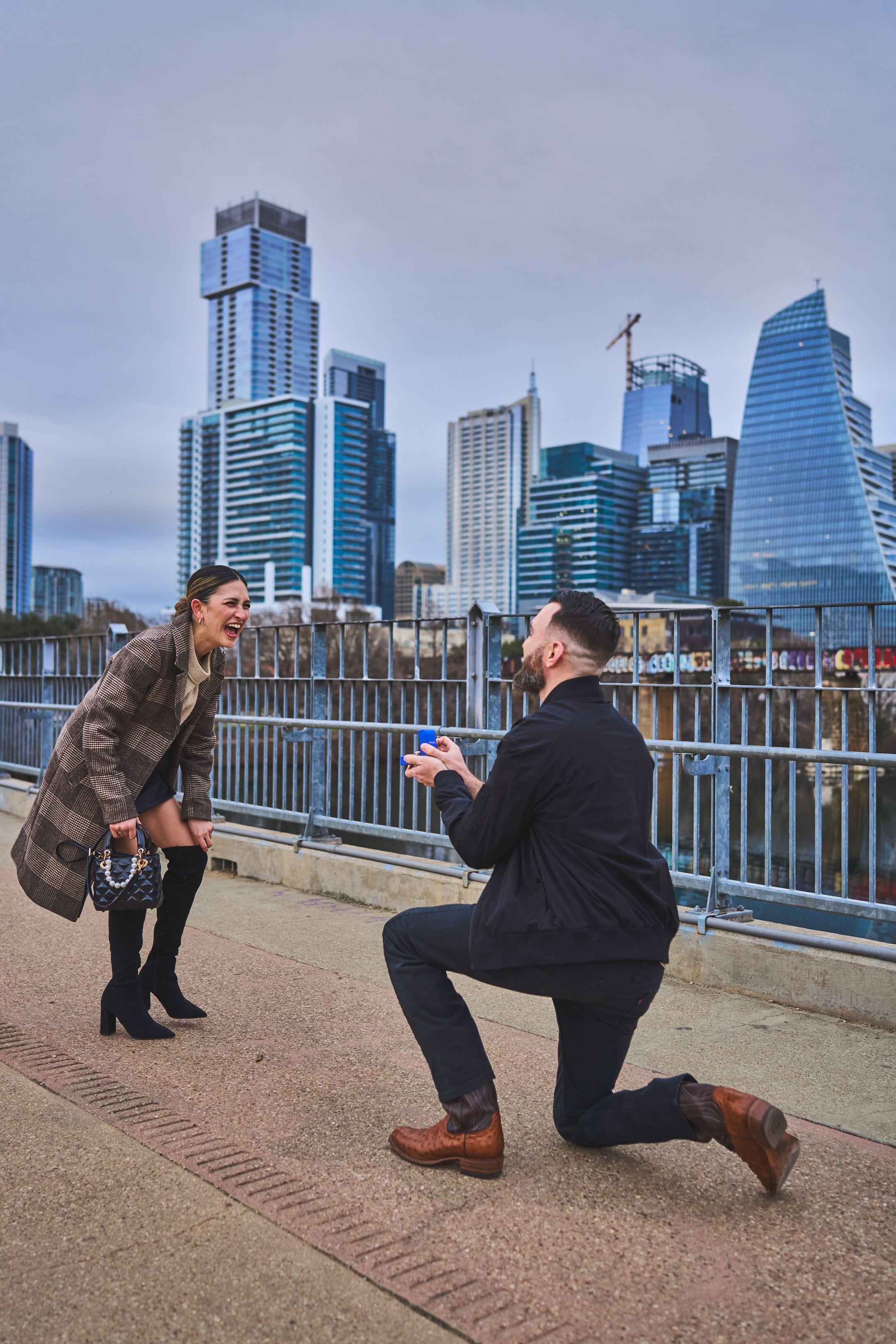 Man on one knee holding out a ring box as his partner reacts, downtown Austin behind them — Engagement & Proposals photography, Austin A man on one knee holding out a ring box as his partner reacts, downtown Austin behind them.