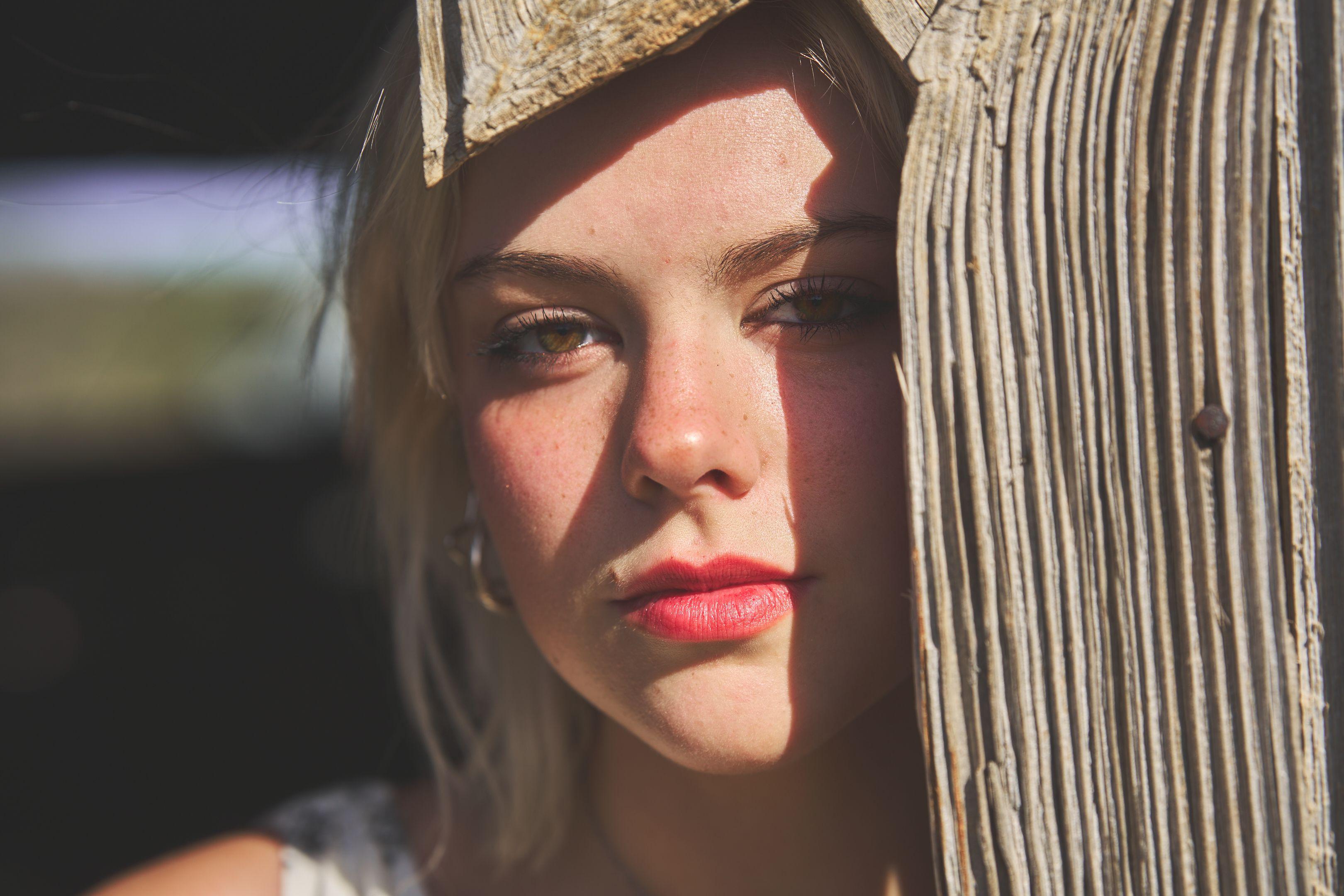 Close portrait of a woman's face with hard dappled light falling across it — Commons Ford Ranch photography, Austin A close portrait of a woman's face with hard dappled light falling across it, beside a weathered wooden post.