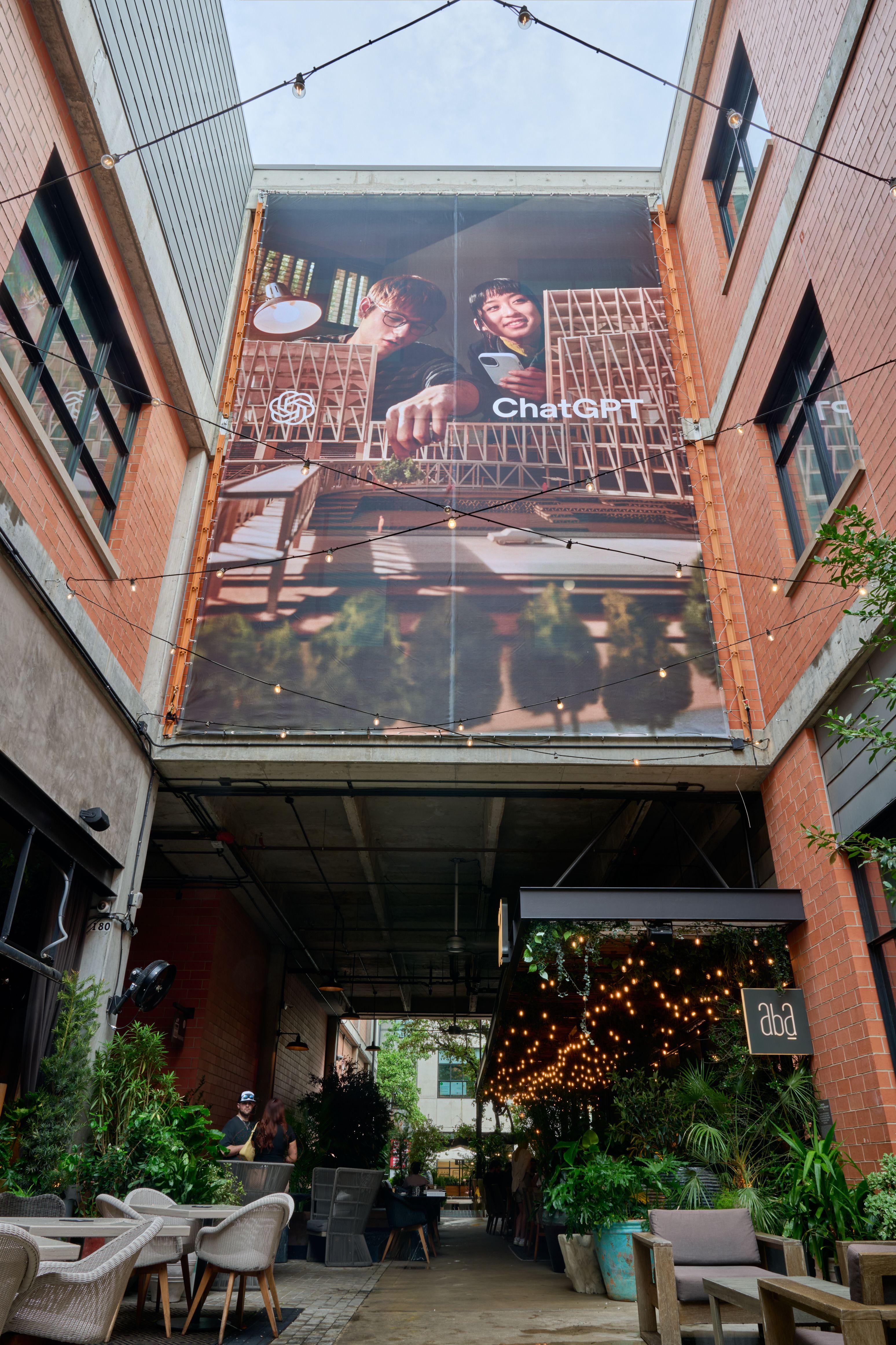 Large ChatGPT billboard mounted above a plant-filled brick alley — Outdoor media photography, Austin A large ChatGPT billboard mounted above a plant-filled brick alley.