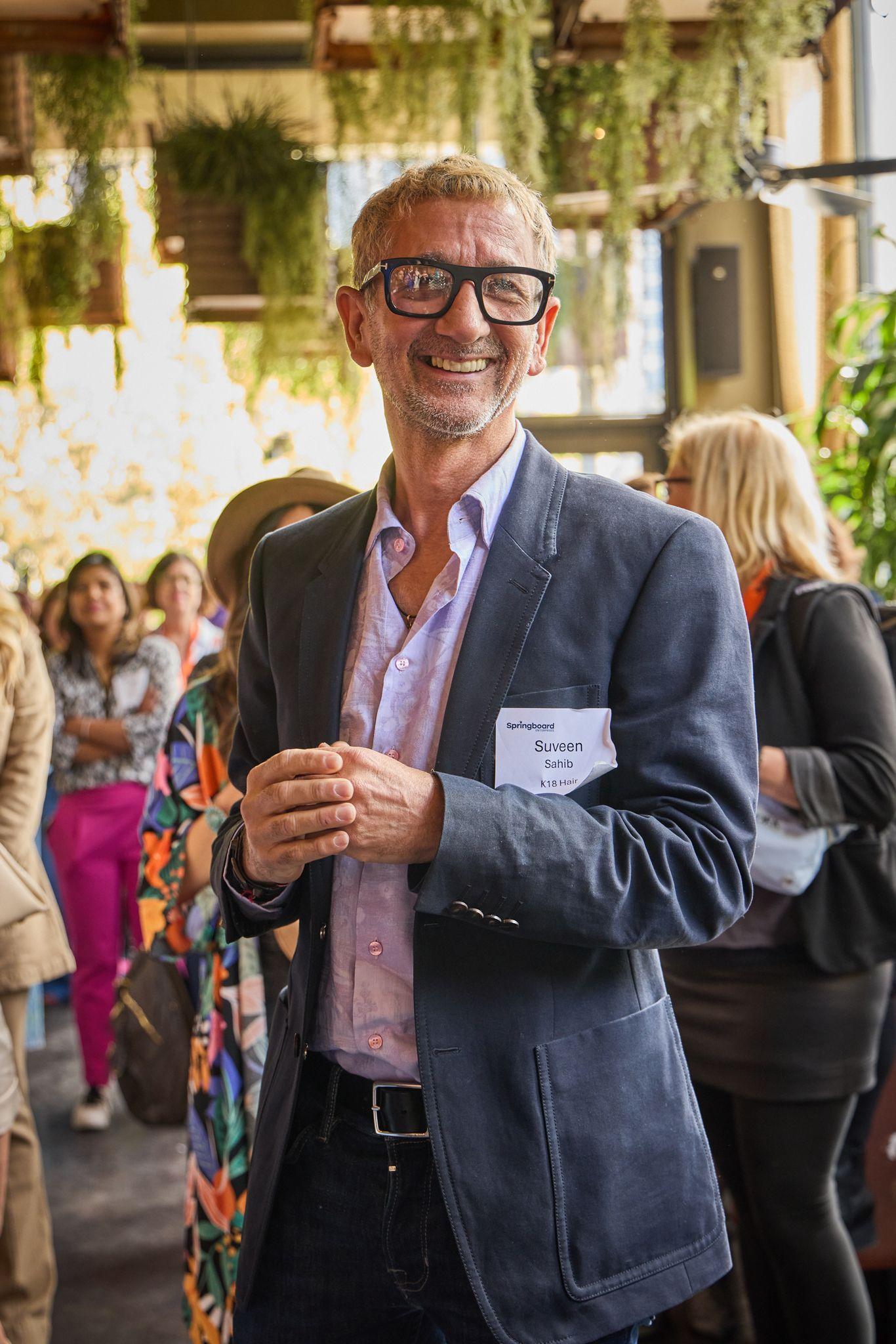 Man in a blazer and name badge smiling in a crowded room at a networking event — Team offsite photography, Austin A man in a blazer and name badge smiling in a crowded room at a networking event