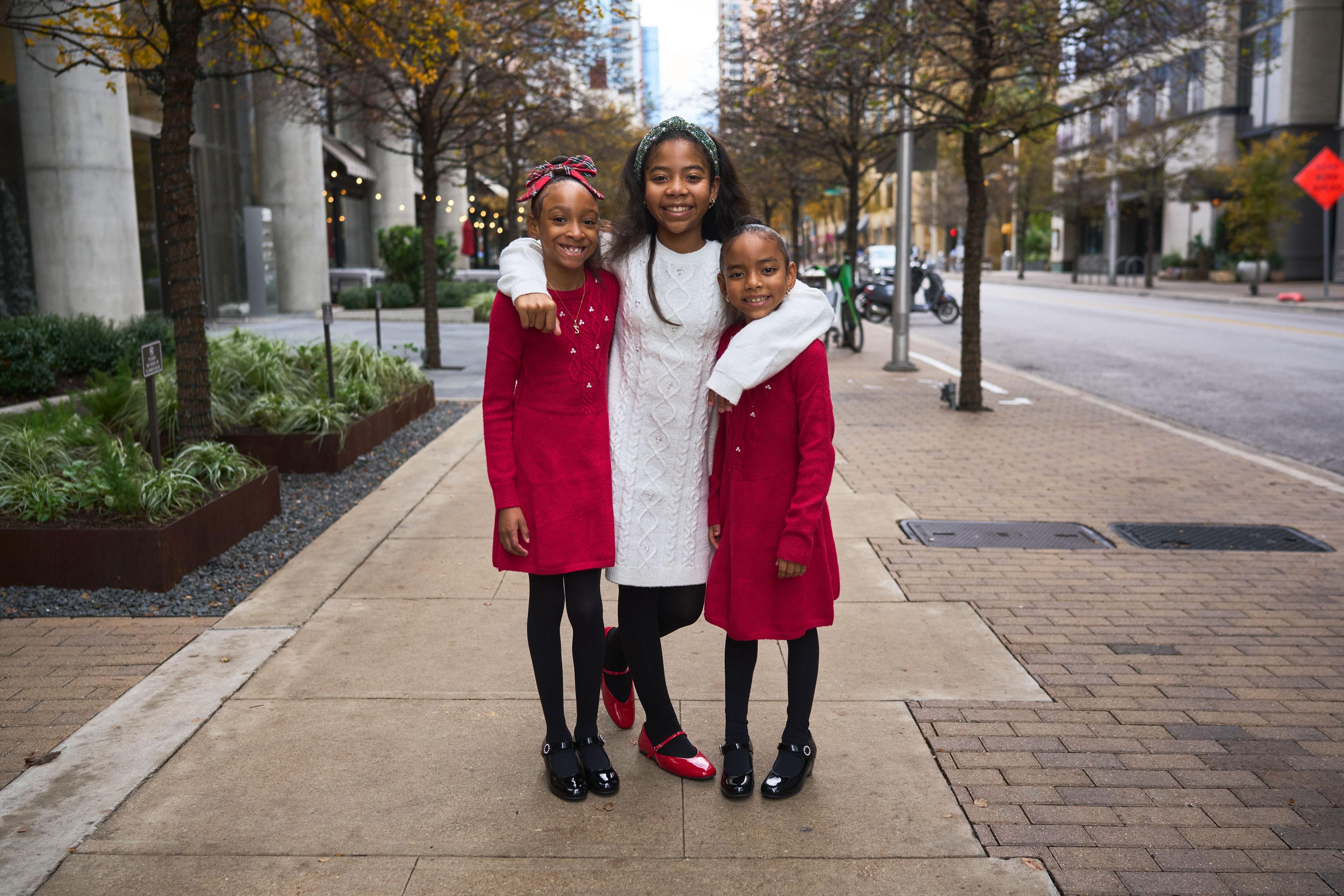 Three girls in red and white coats standing on a tree-lined downtown pavement — Downtown Austin photography, Austin Three girls in red and white coats standing on a tree-lined downtown pavement.