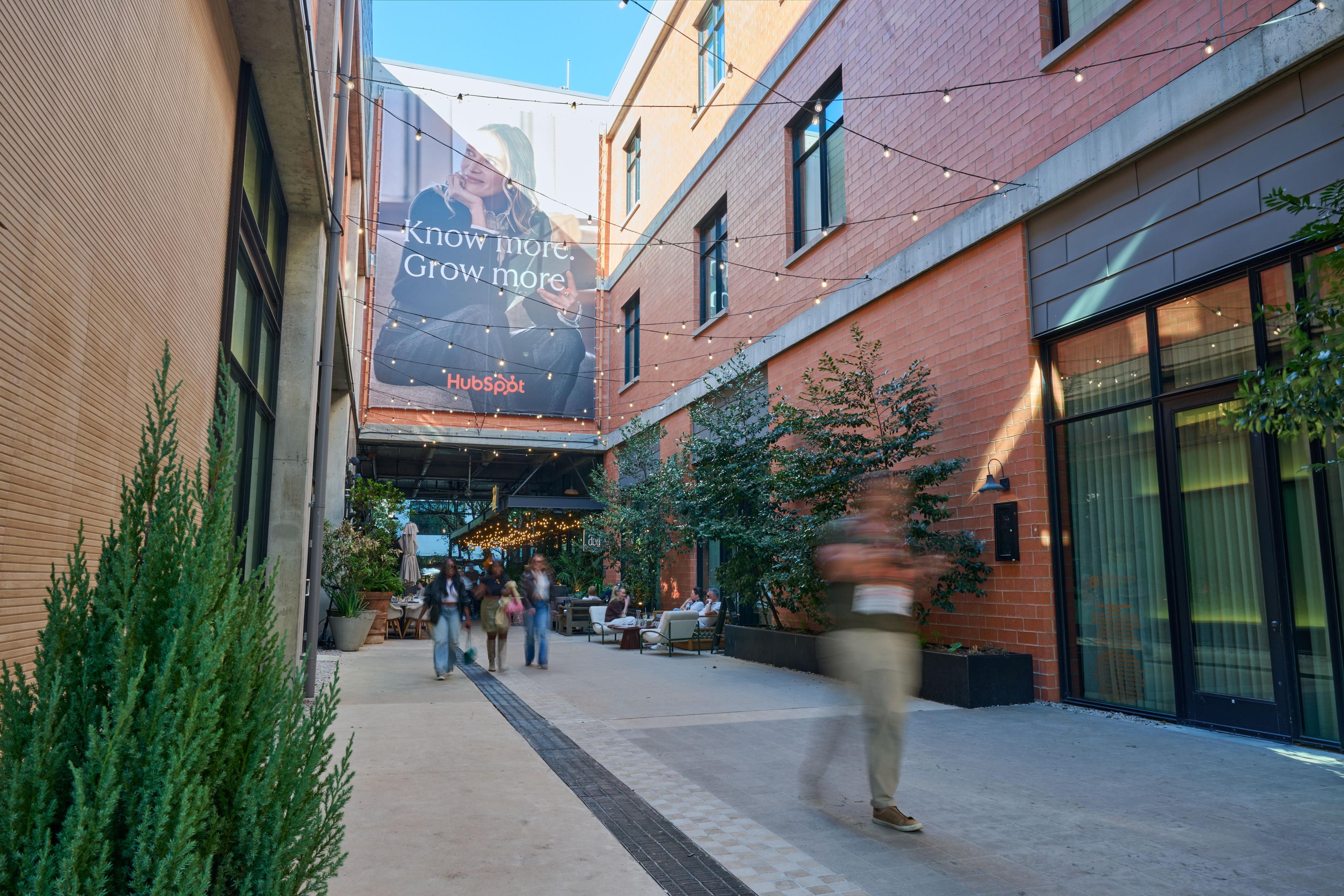 HubSpot 'Know more. Grow more.' wall banner in a brick pedestrian alley — Outdoor media photography, Austin A HubSpot 'Know more. Grow more.' wall banner in a brick pedestrian alley with people walking past.