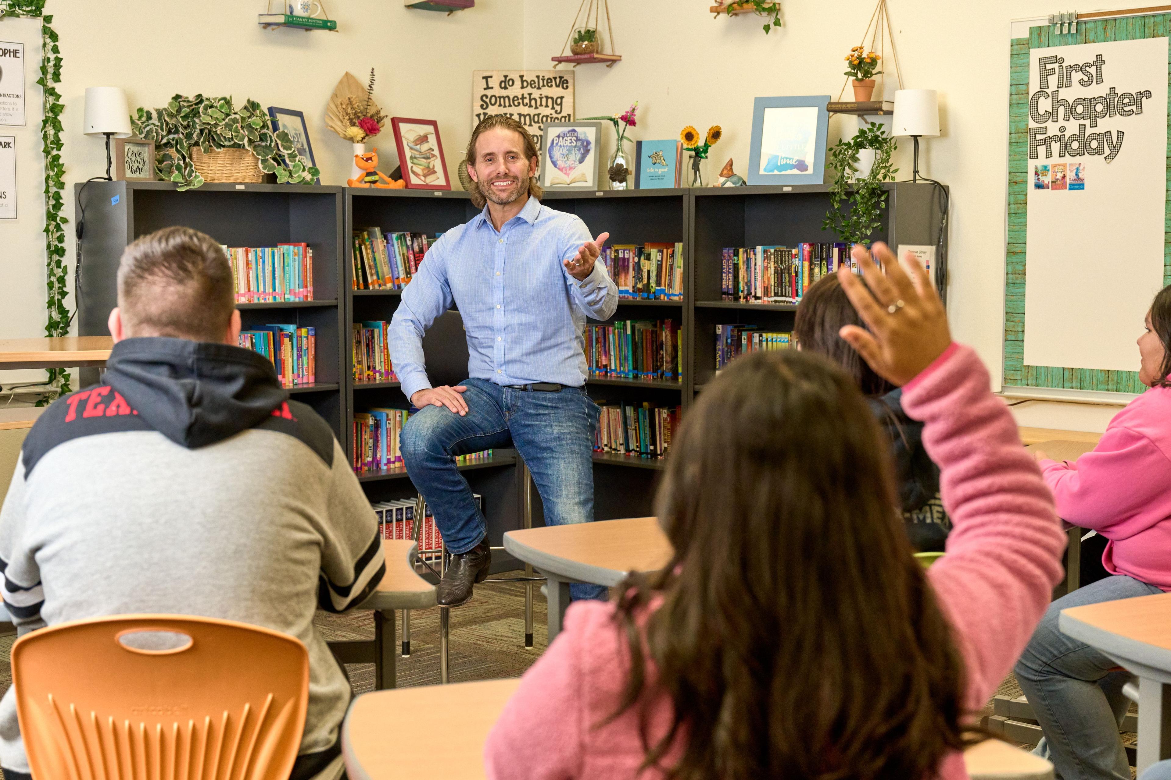 Man seated in a school library talking to students, books on the shelves behind him — School event photography, Austin Man seated in a school library talking to students, books on the shelves behind him